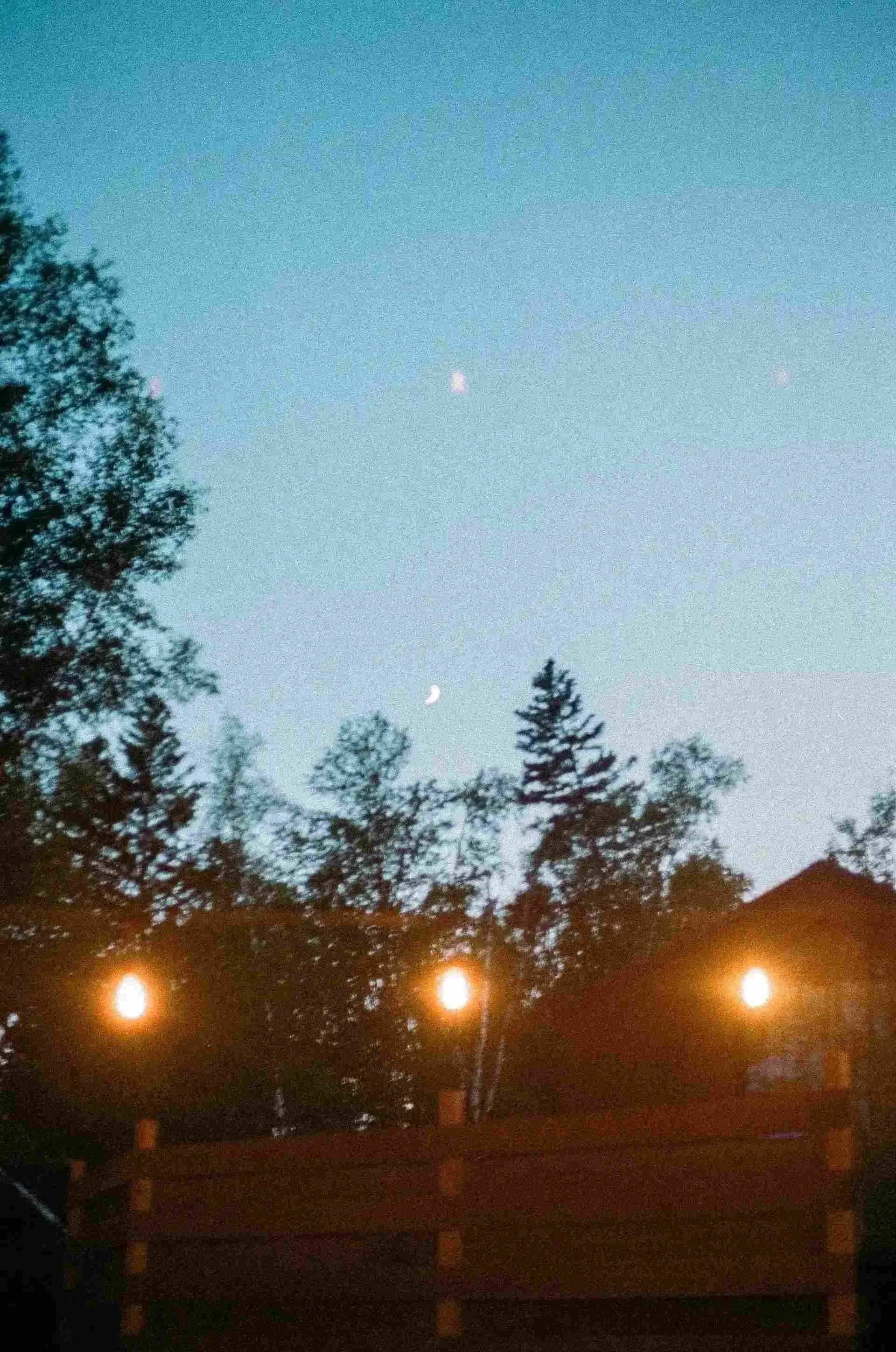 Night sky with crescent moon and scattered lights above silhouetted trees and rooftop.