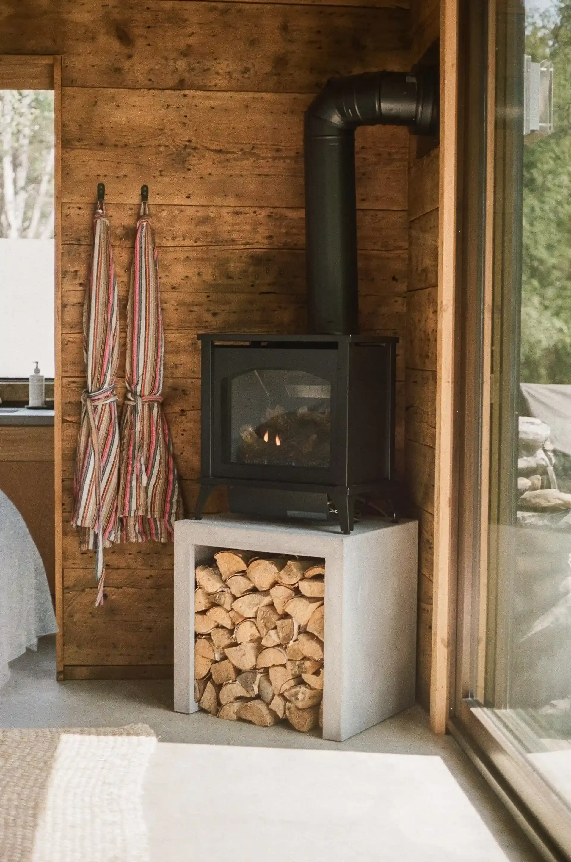 Cozy indoor scene with a wood-burning stove on a concrete base filled with firewood, adjacent to a wooden wall with two striped towels hanging.