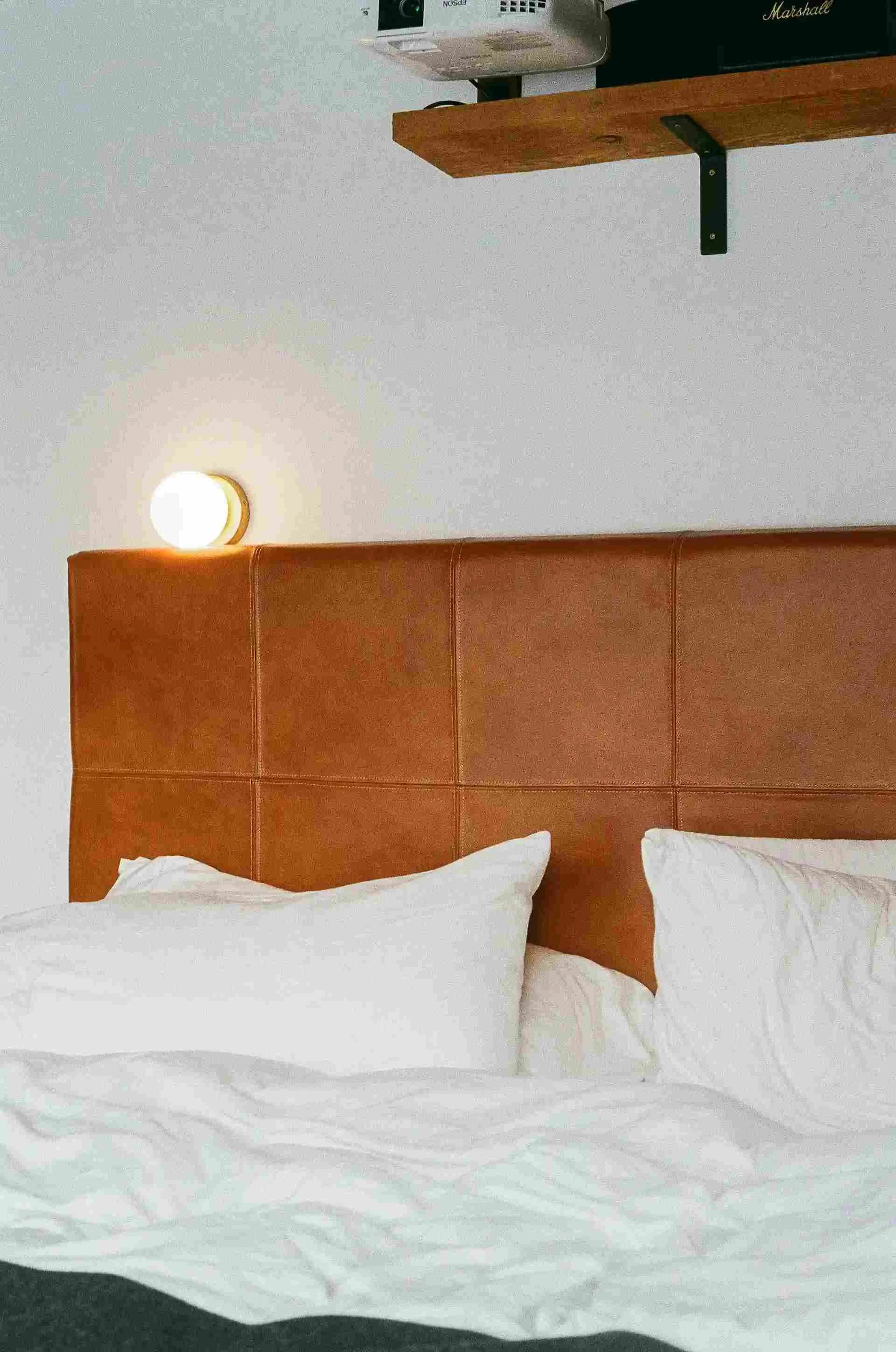 Modern bedroom with a brown leather headboard, white bedding, a wall-mounted round light, and a wooden shelf holding a projector and speaker.