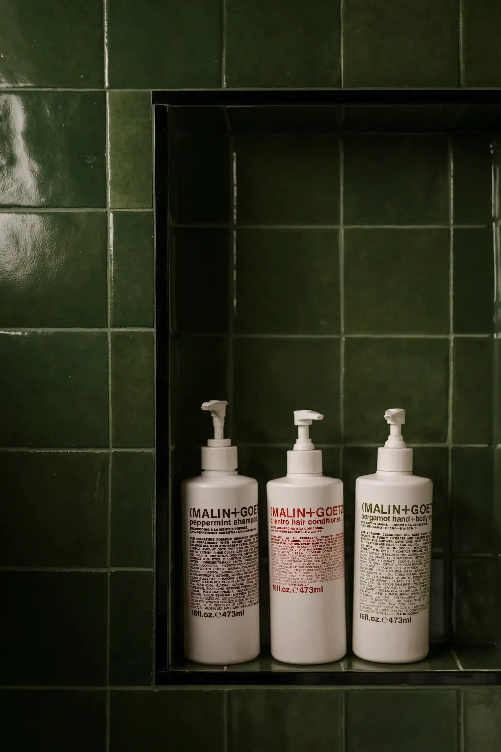 Three Malin+Goetz bottles on a green tiled shelf.