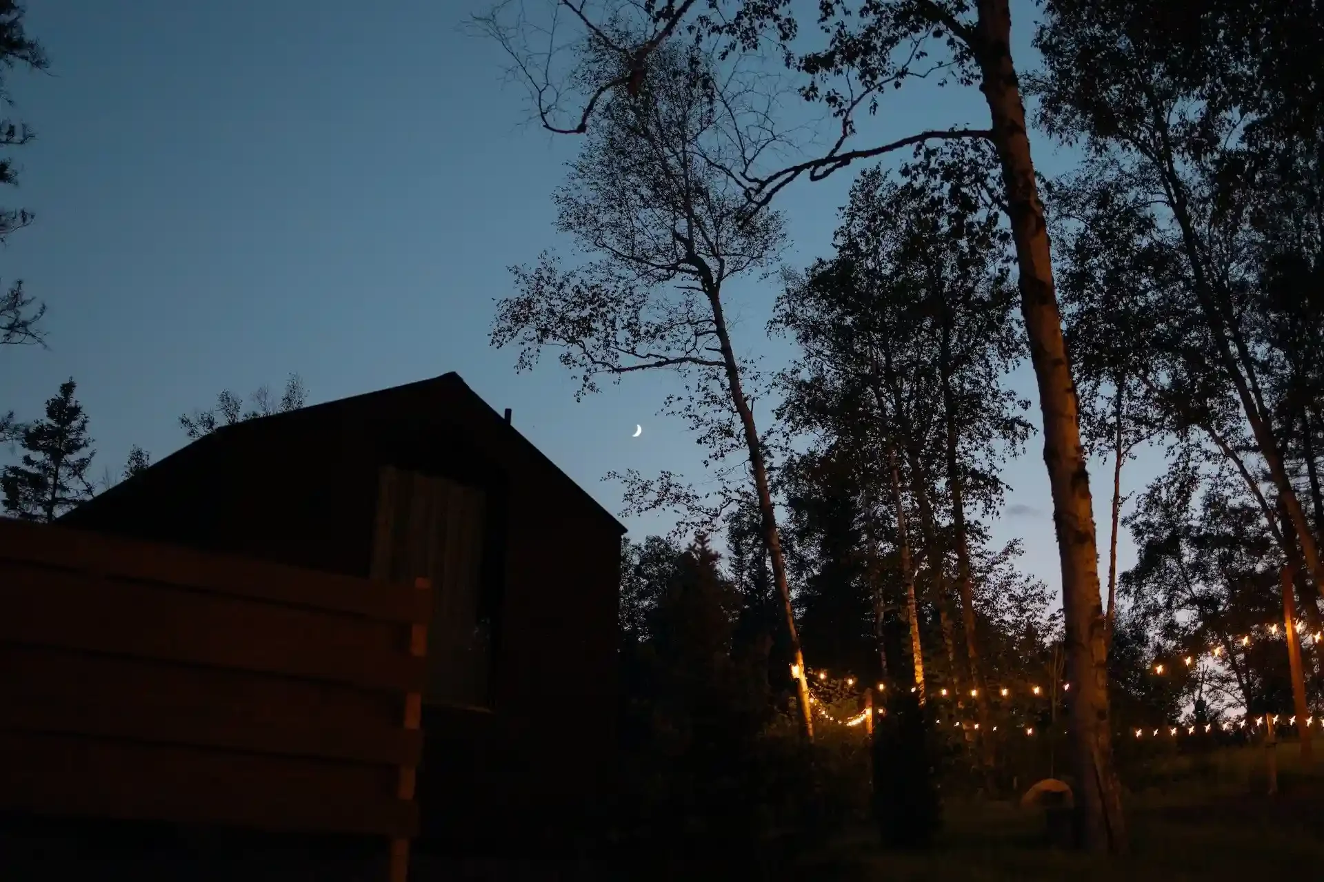Night scene with a cabin silhouette, trees, crescent moon, and string lights in a woodland setting.