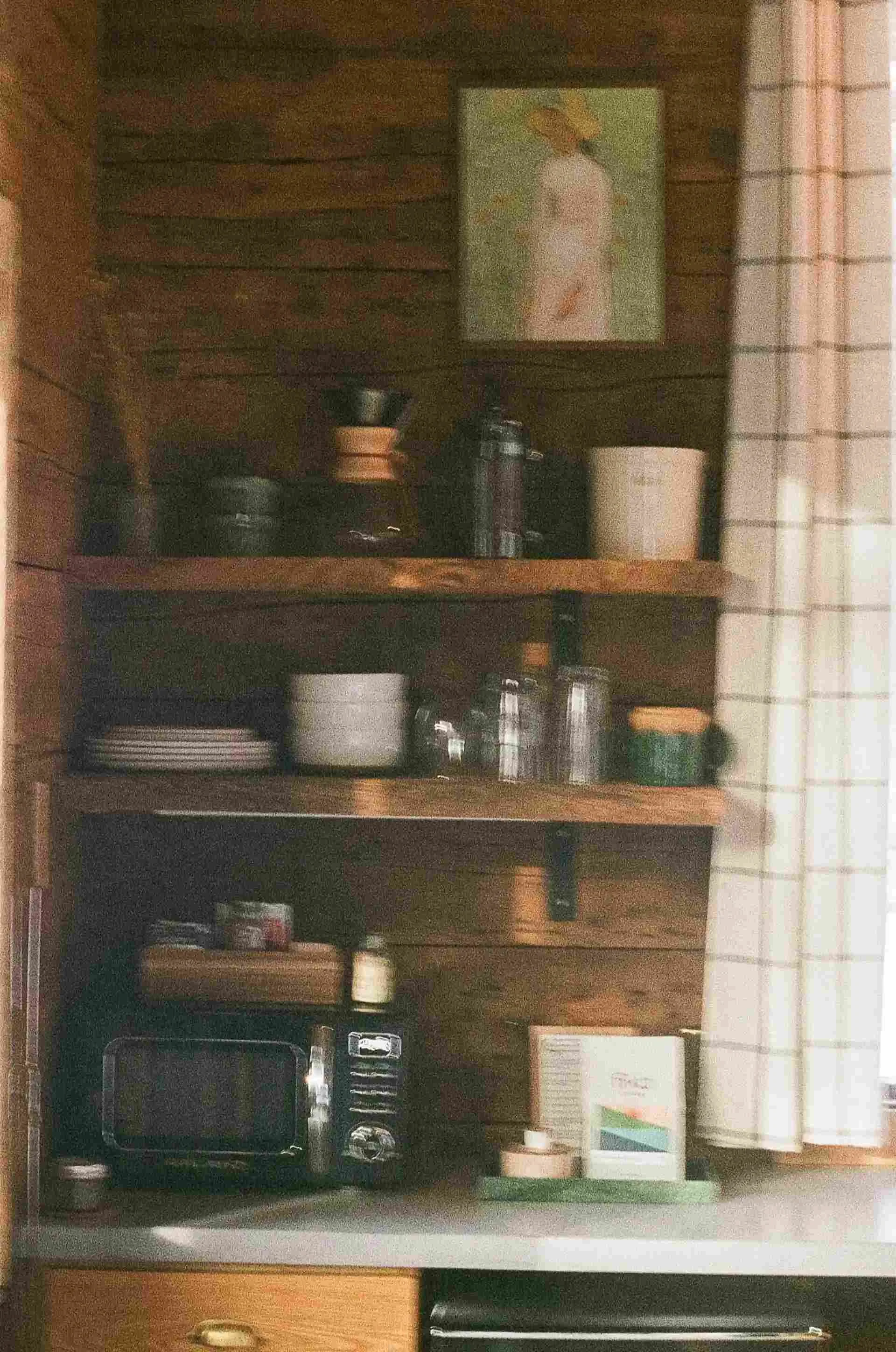 Rustic kitchen with wooden shelves, a painting, kitchenware, a coffee maker, glass jars, a toaster oven, and a curtain by the window.