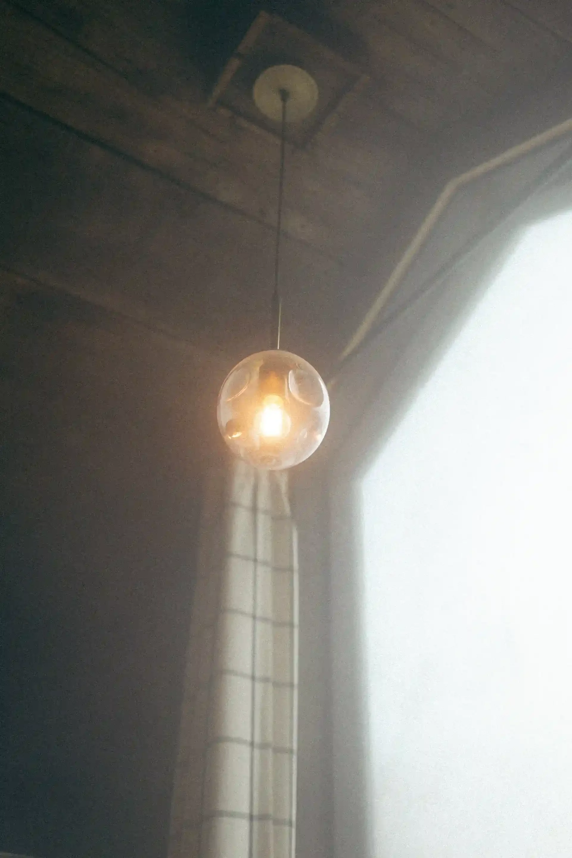 A hanging spherical pendant light with a warm glow inside a room with wooden ceiling and sheer curtains.
