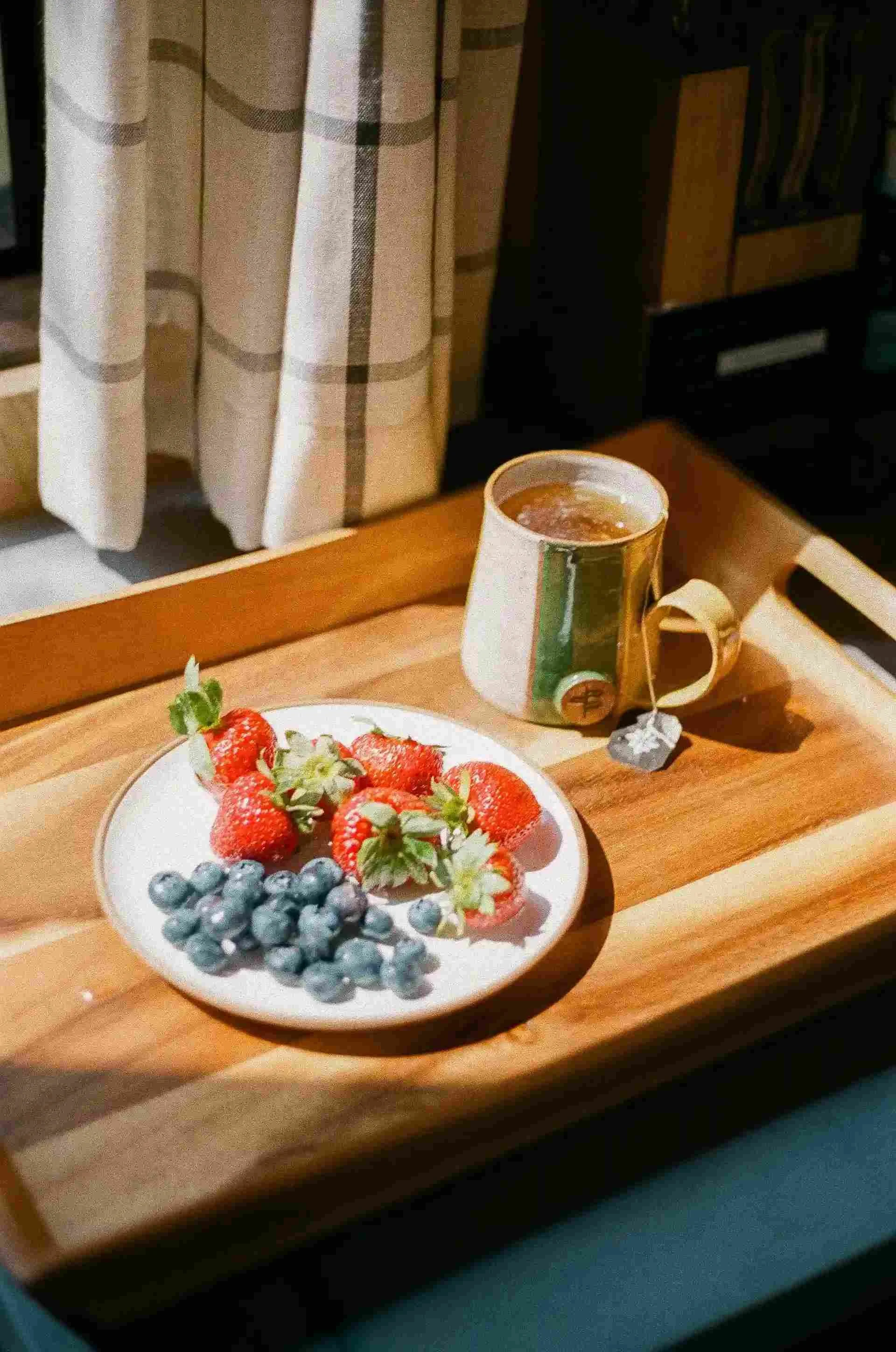 A wooden tray with a plate of strawberries and blueberries alongside a mug of tea, near a window with plaid curtains.