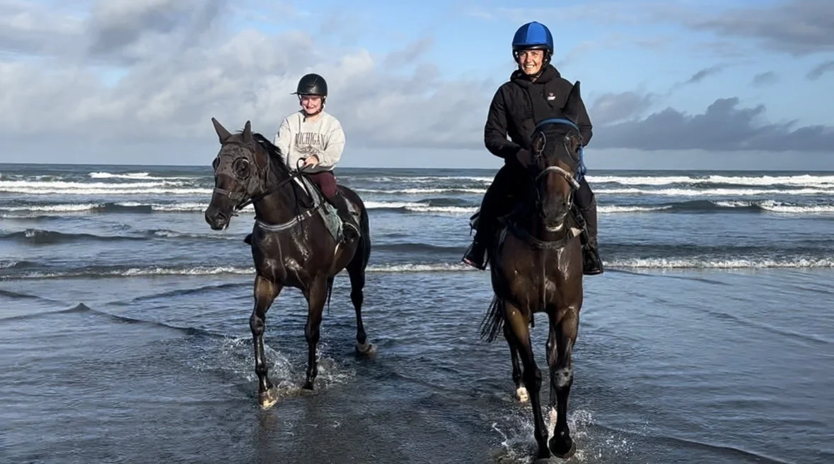 Two people riding horses in the shallow ocean water on a cloudy day.