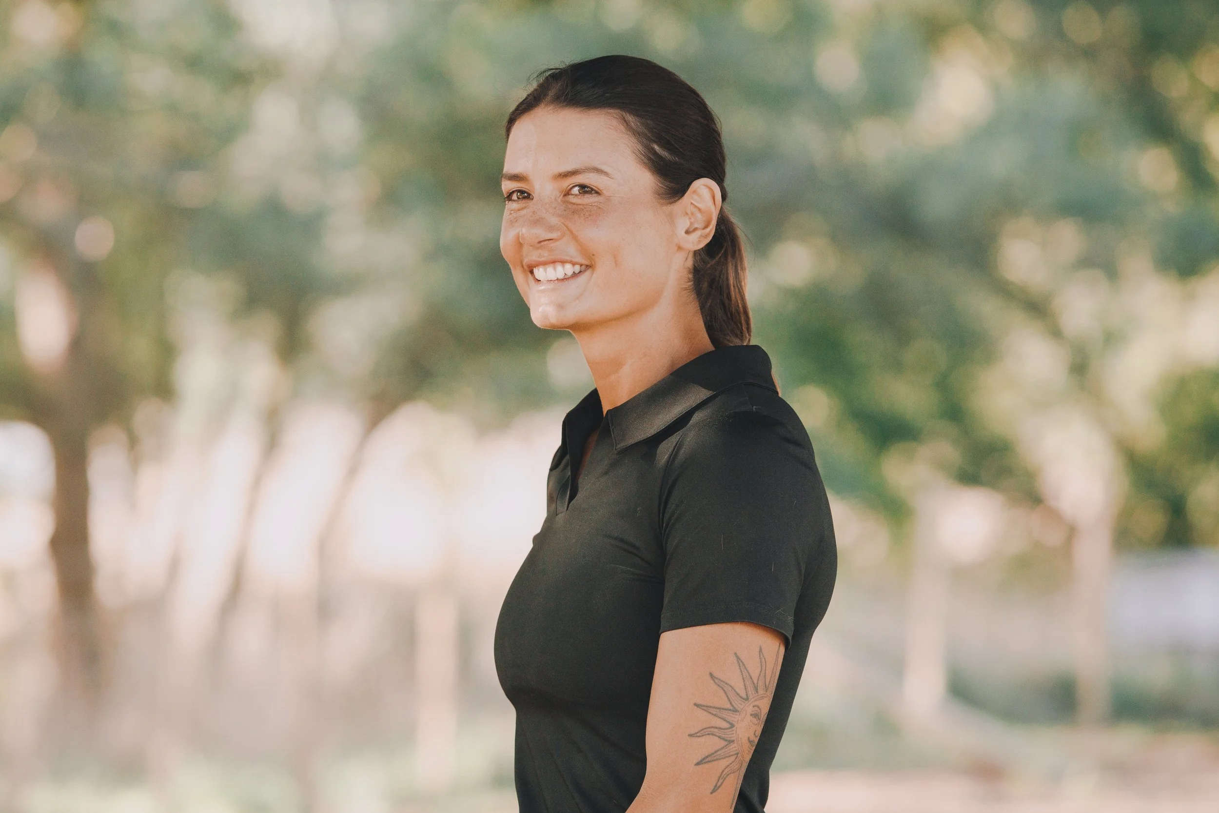 Smiling woman with dark hair tied back, wearing a black polo shirt with a sun tattoo on her arm, outdoors with trees in the background.