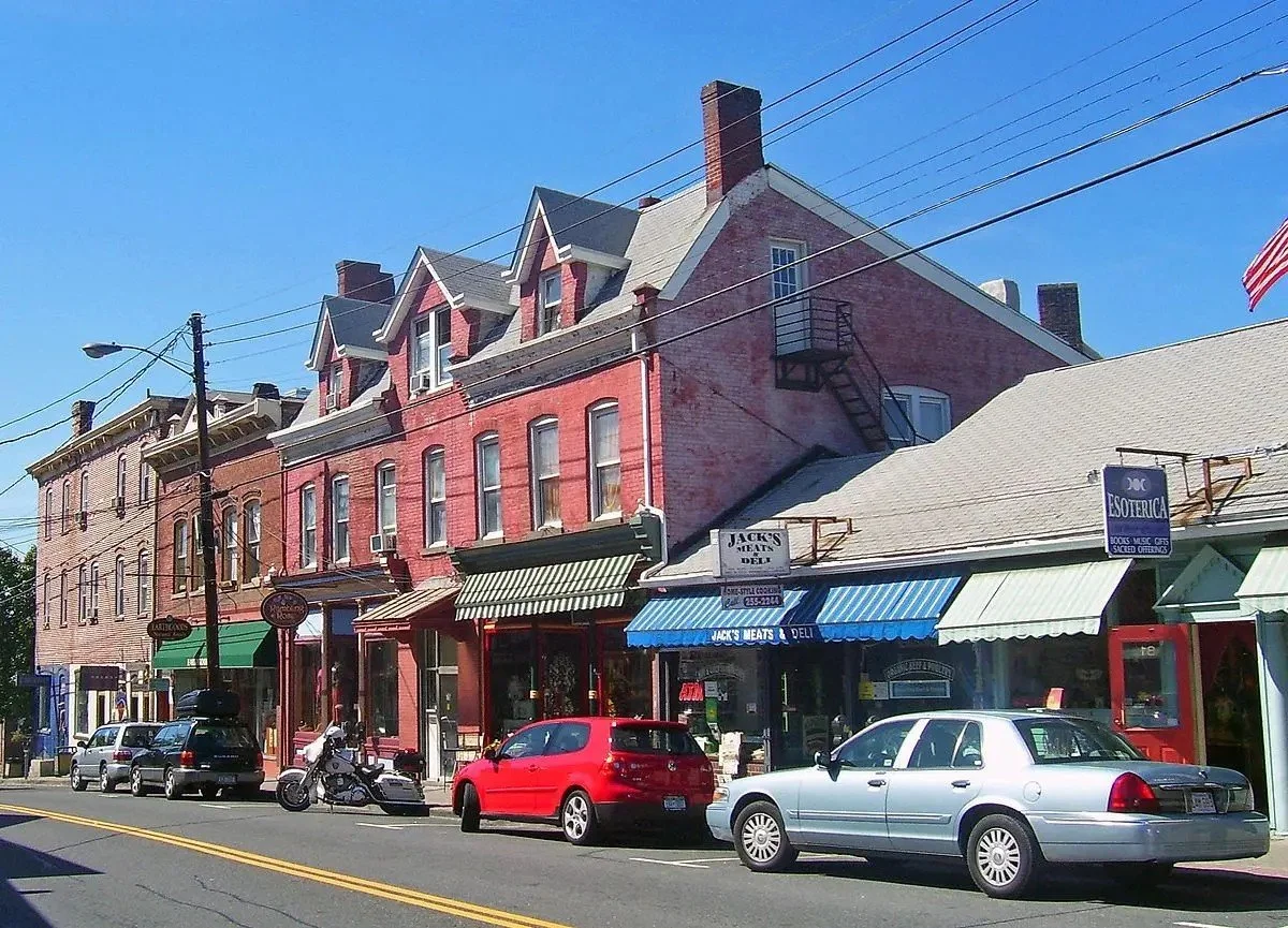A street view with brick buildings, parked cars, and storefronts including Jack's Meats and Deli with blue awning, and a sign for Esoteric books and gifts. Clear sky.