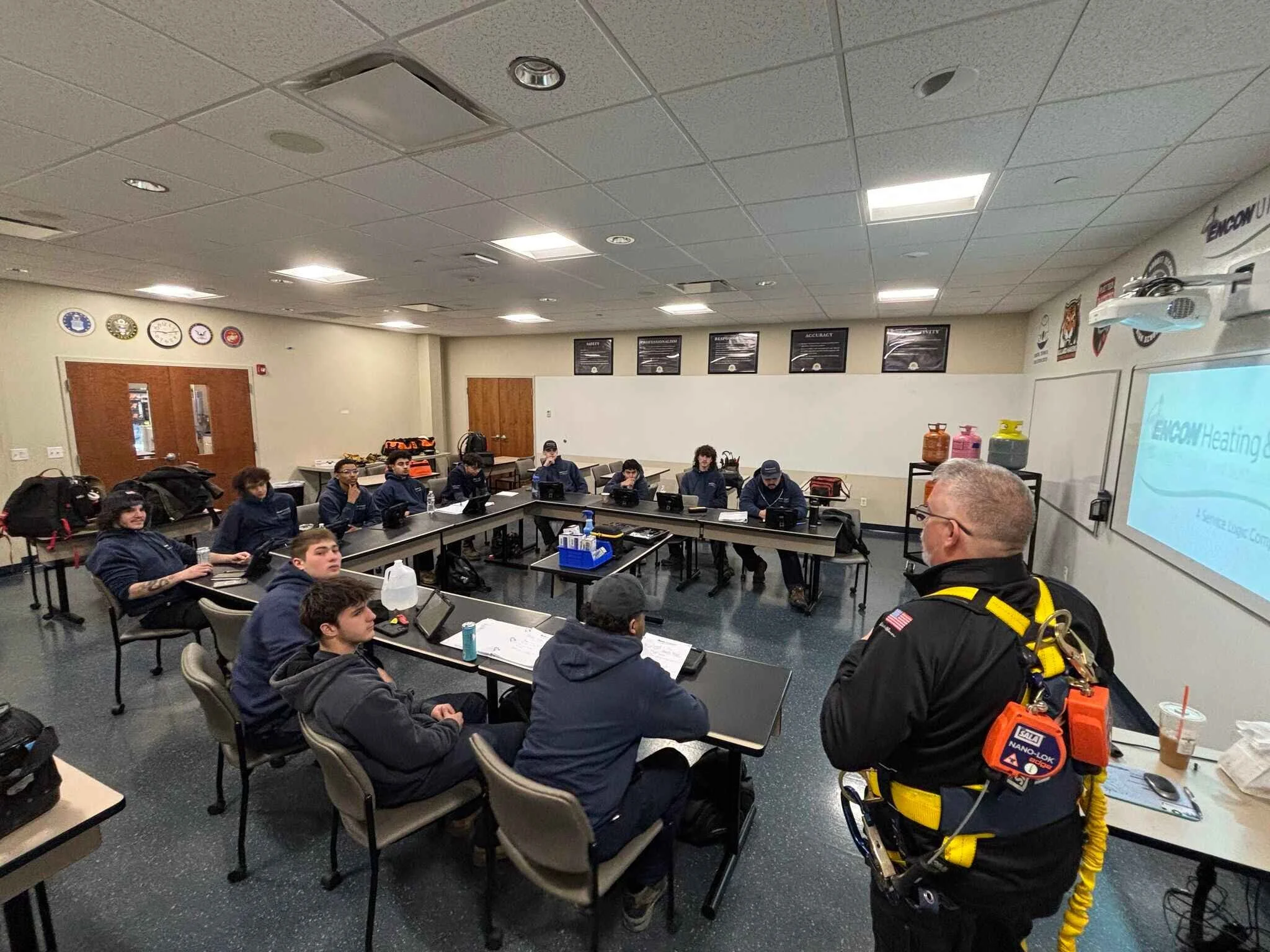 A classroom filled with students sitting at tables, listening to a man in safety gear who is giving a presentation, with a whiteboard and projection screen displaying a logo.