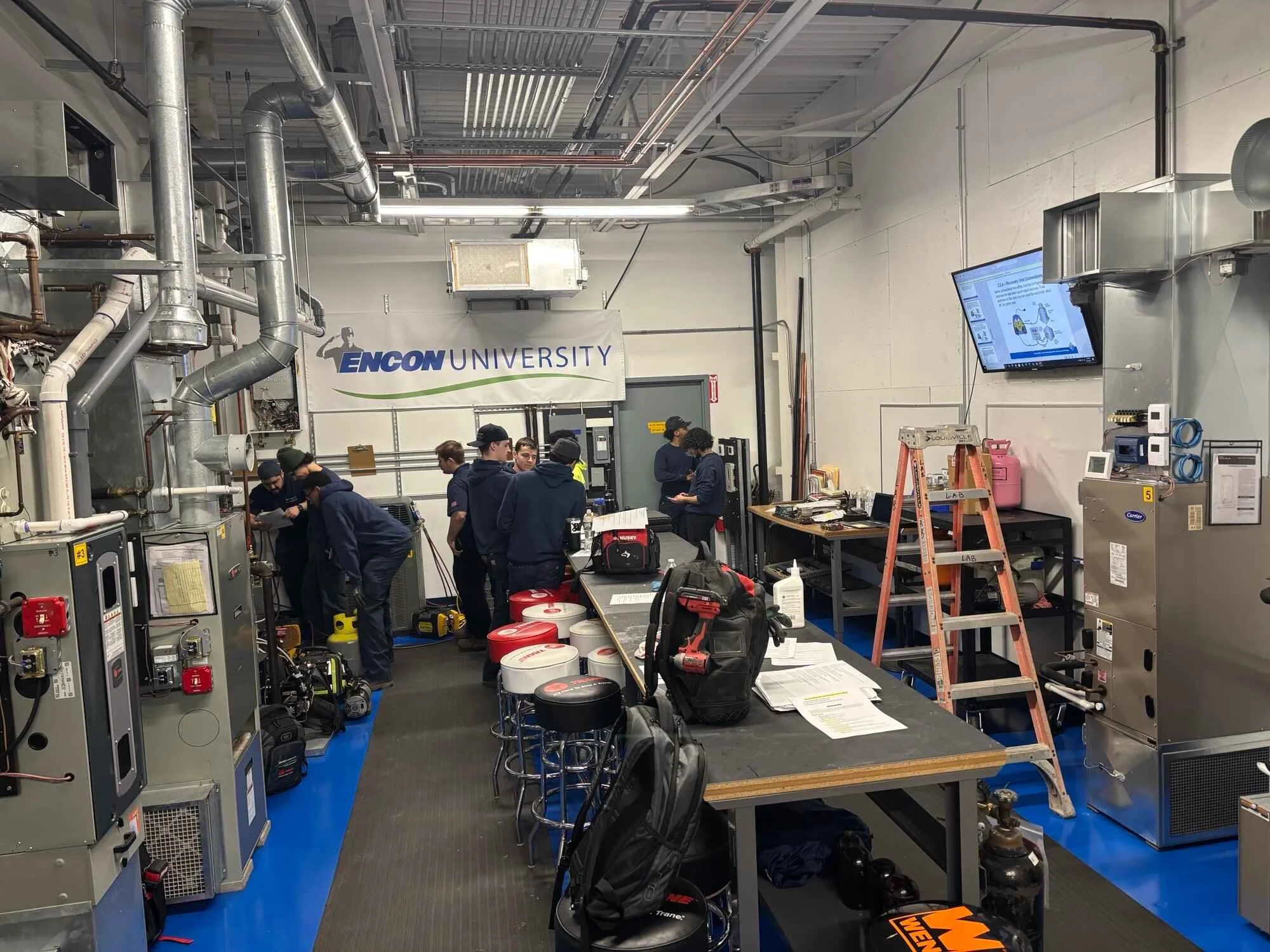 Group of students working in a laboratory at Encon University. The lab has industrial equipment, tools, and a large screen displaying a presentation.