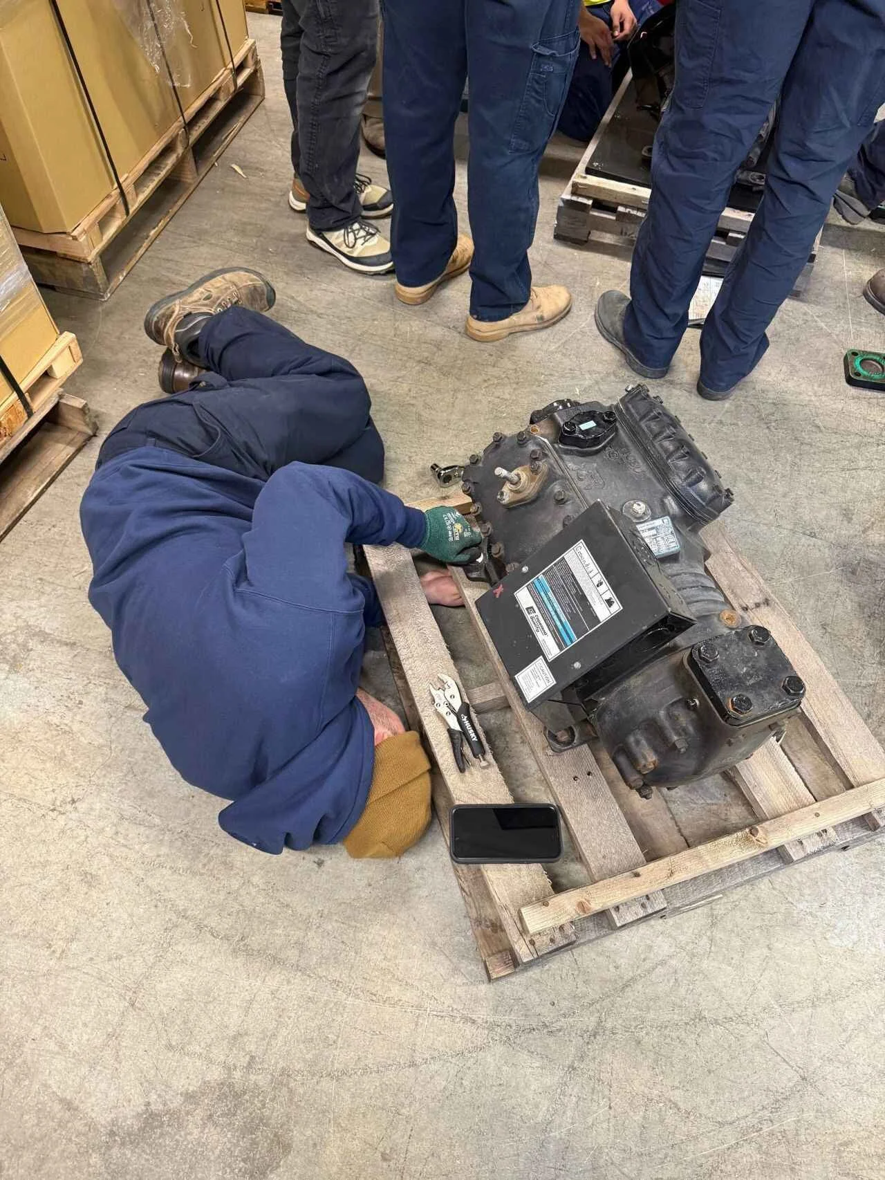 A worker fixing an engine on a wooden pallet in a warehouse, surrounded by several people.