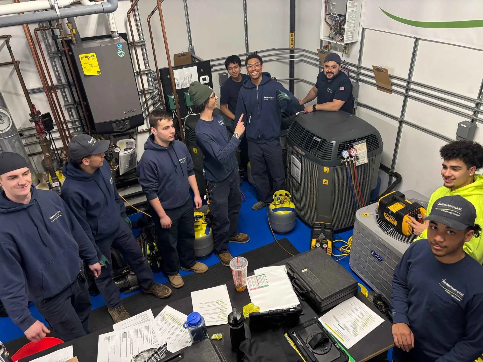 Group of people in HVAC training room with equipment and tools, engaged in a classroom or hands-on session.