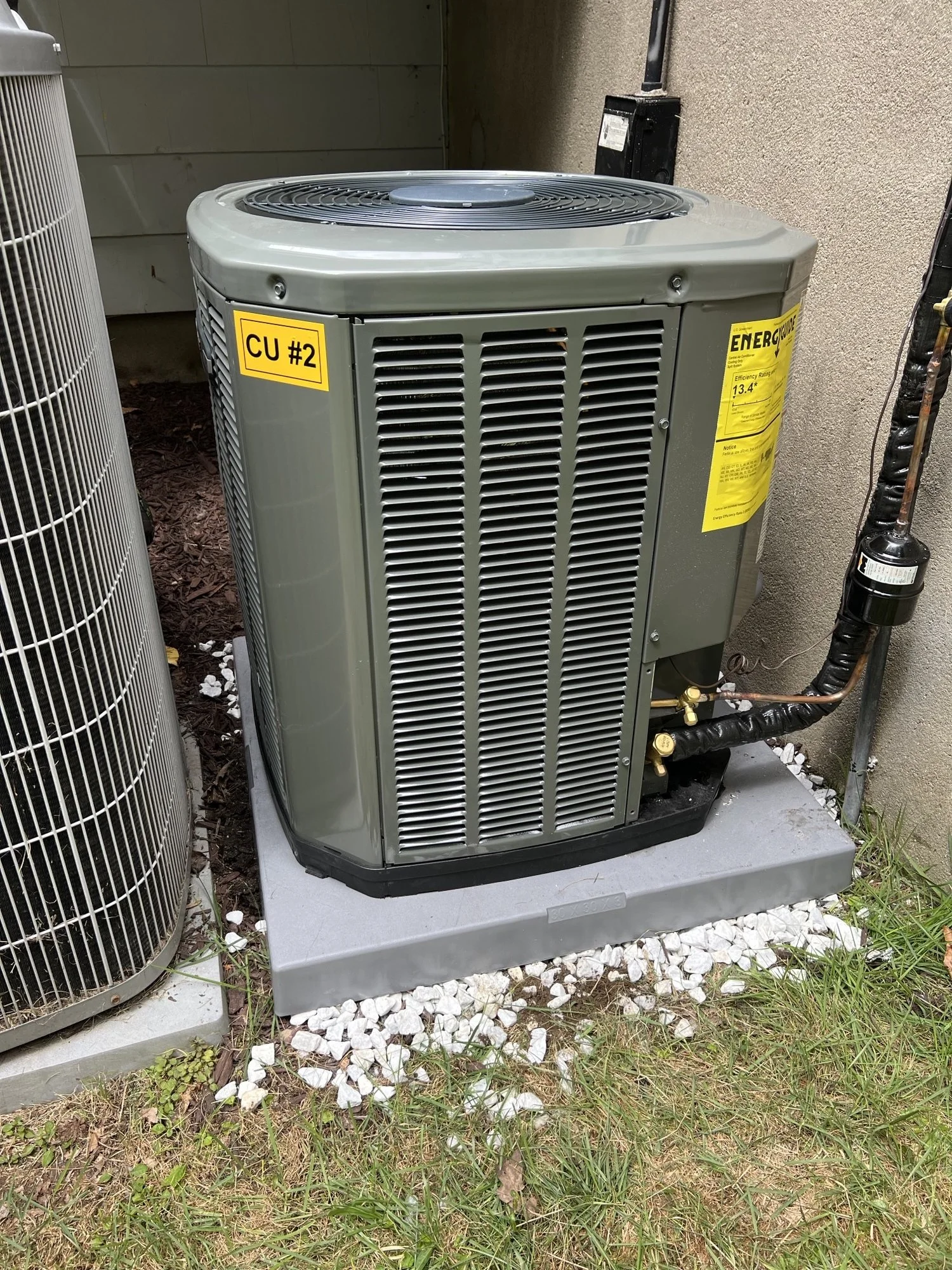 An outdoor HVAC unit installed on a concrete pad next to a beige wall, with a label reading 'CU #2' and an energy efficiency sticker.