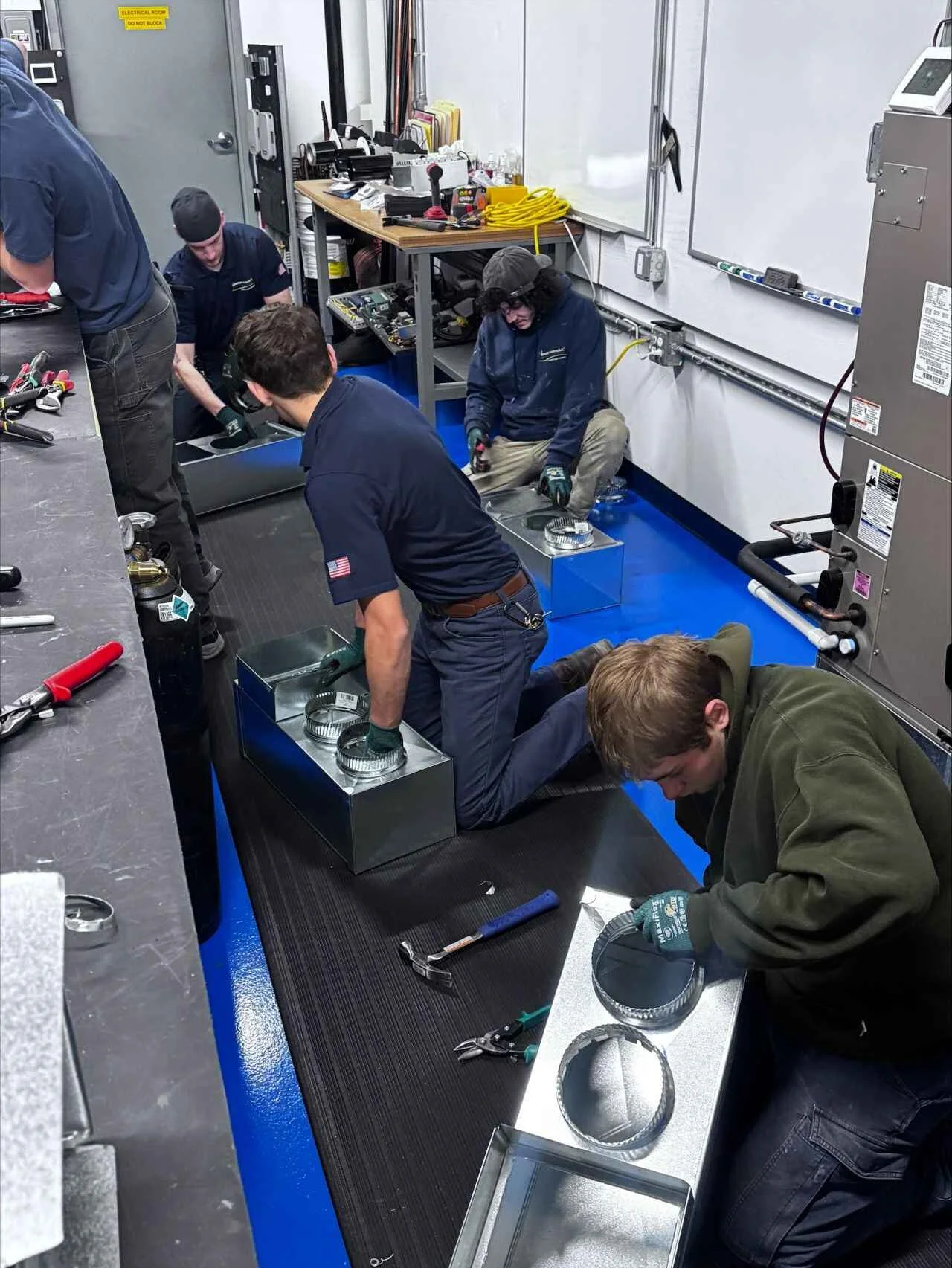 Group of five people working with metal components in an industrial workshop, using tools and wearing gloves, with various equipment and tools around.