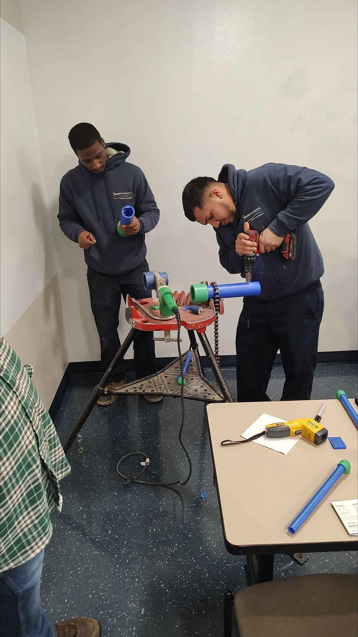 Two men working on PVC pipe project in a room, one using a power drill, the other holding a pipe, with tools on a table nearby.