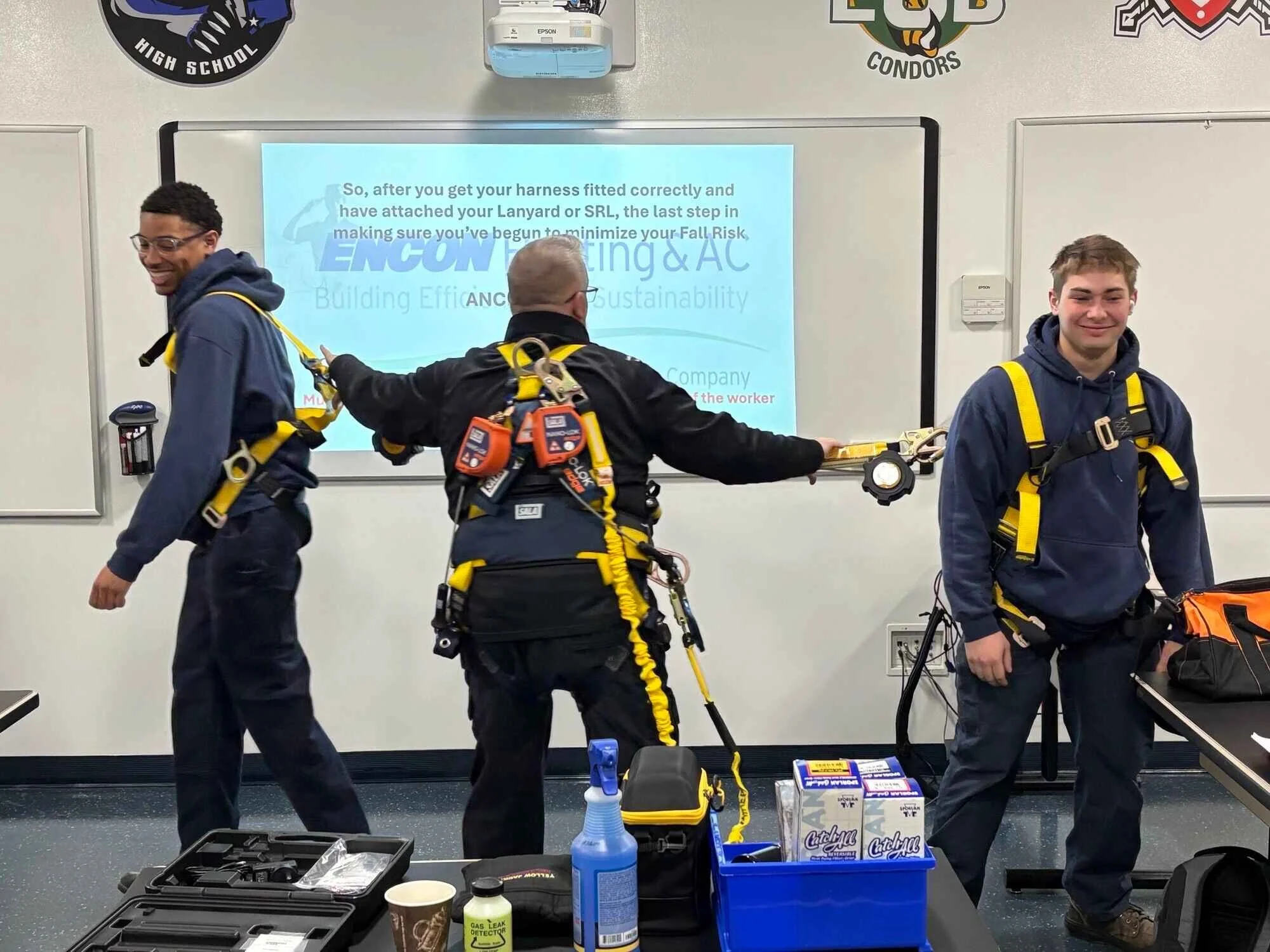 Three students in a classroom wearing safety harnesses connected to a training line, participating in a safety training exercise. Two are smiling, and a projector screen behind them displays instructions about harness fitting.