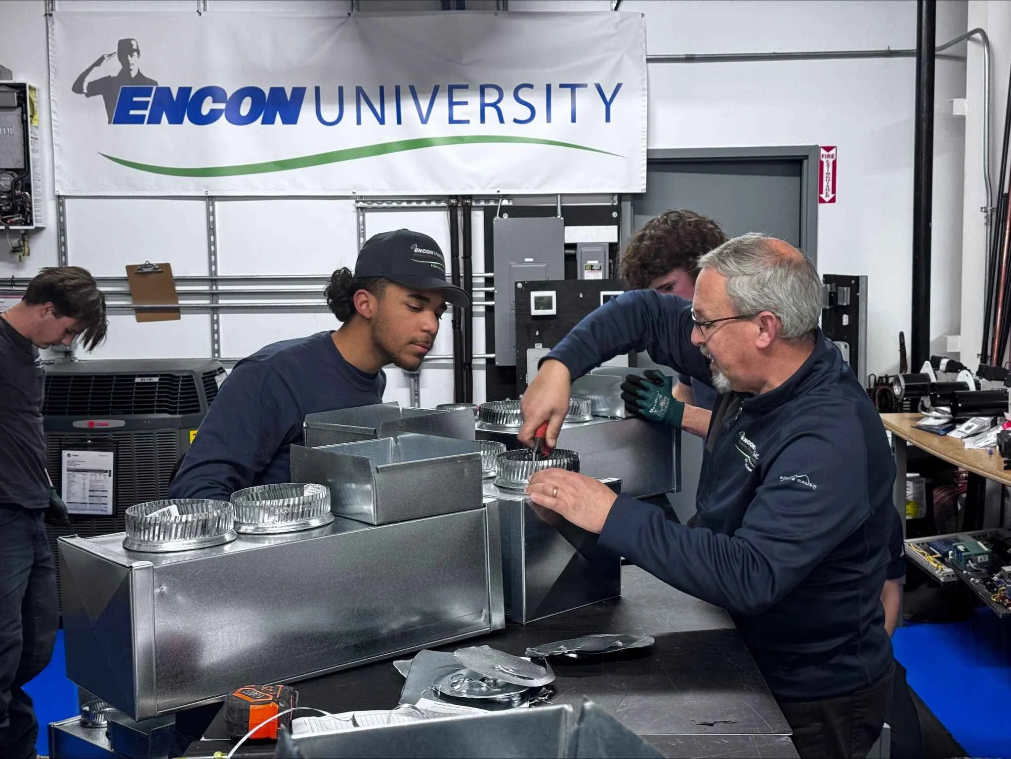 A group of students and an instructor working on a mechanical project in a workshop, with a banner reading 'ENCON UNIVERSITY' in the background.