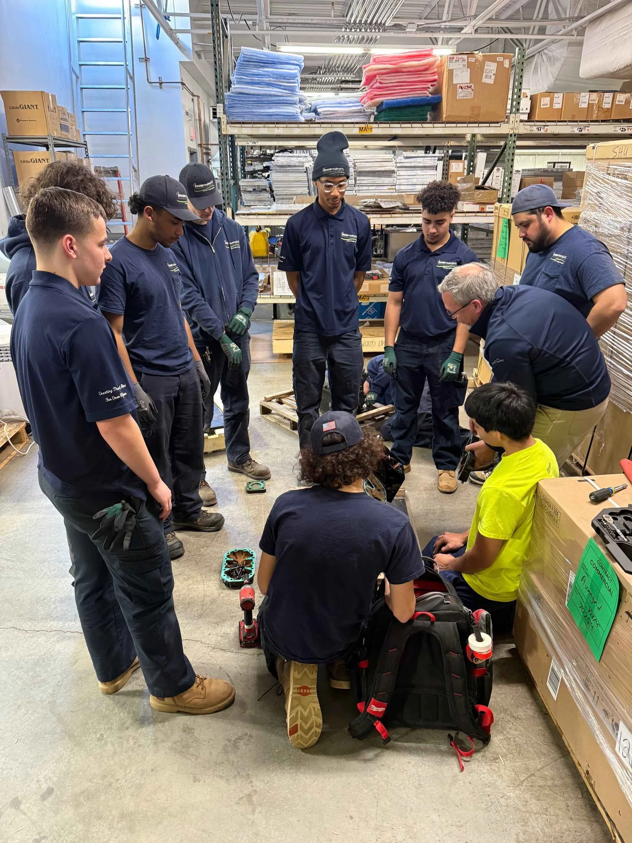 Group of young workers and an instructor gathered in a warehouse, closely inspecting equipment on the floor.