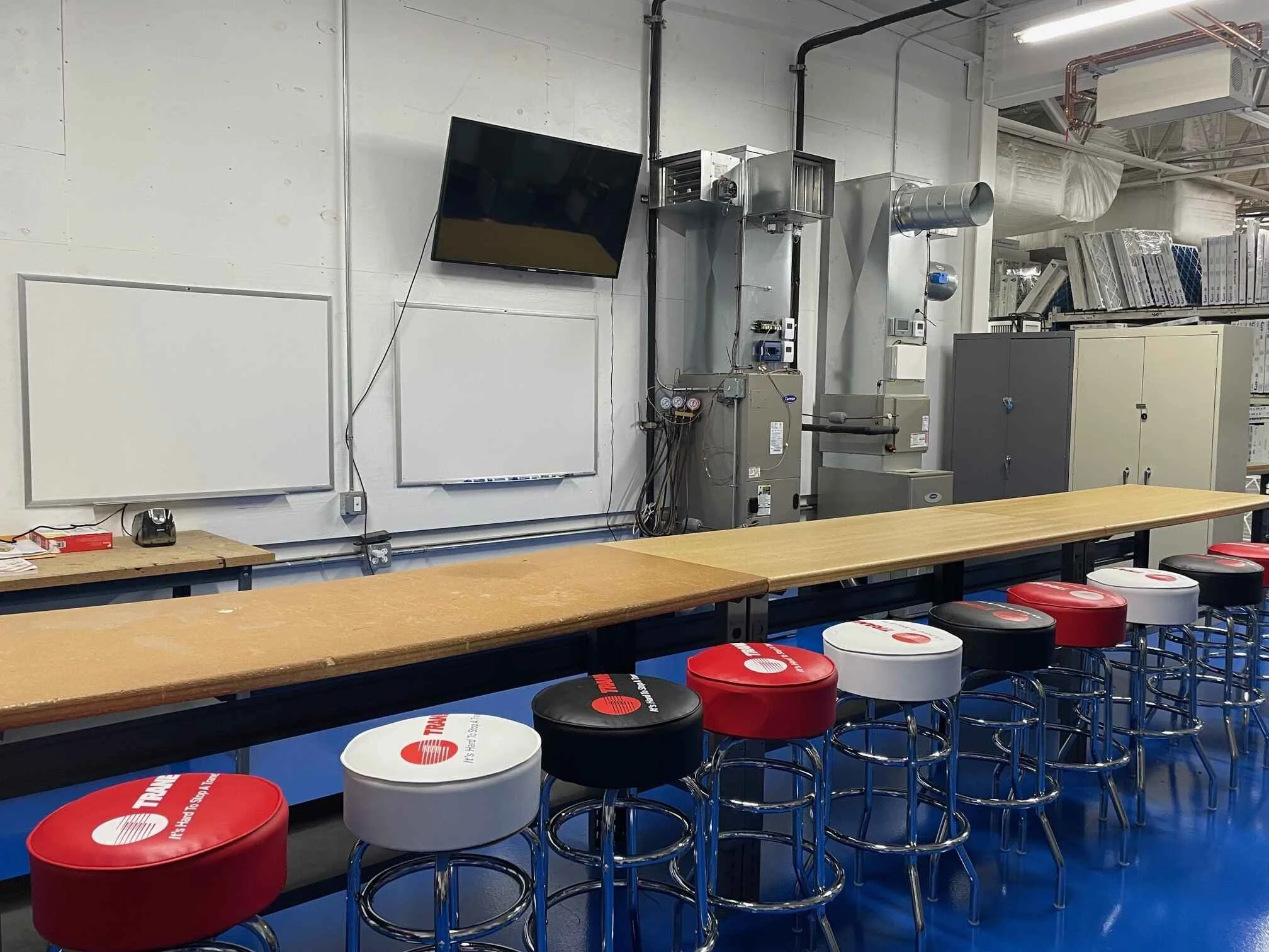 A classroom or training room with a long wooden table, stools with red, white, and black cushioned seats, a flat-screen TV on the wall, and industrial equipment and supplies in the background.
