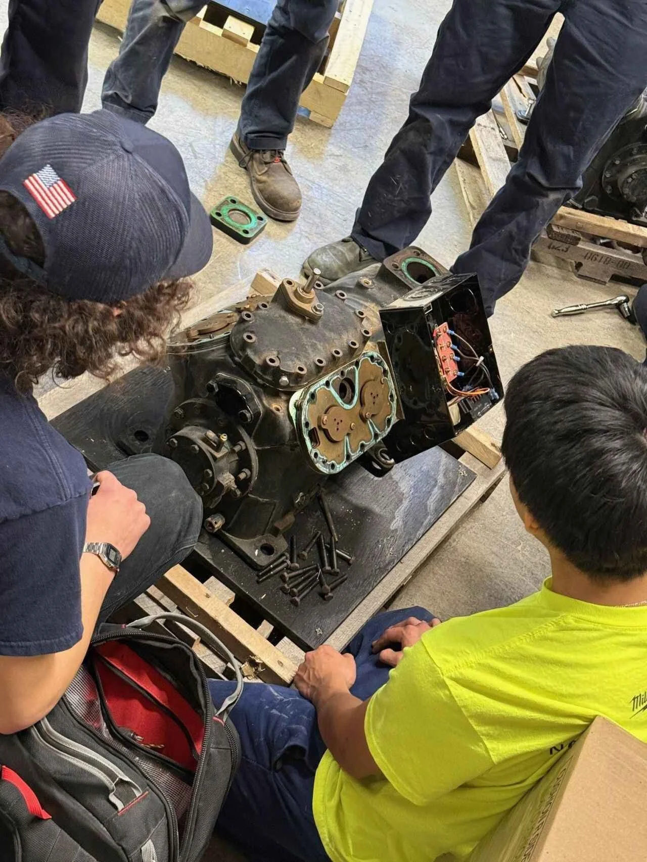 Group of people gathered around a large engine or machine part on a work table, inspecting and assembling it in a workshop or industrial setting.
