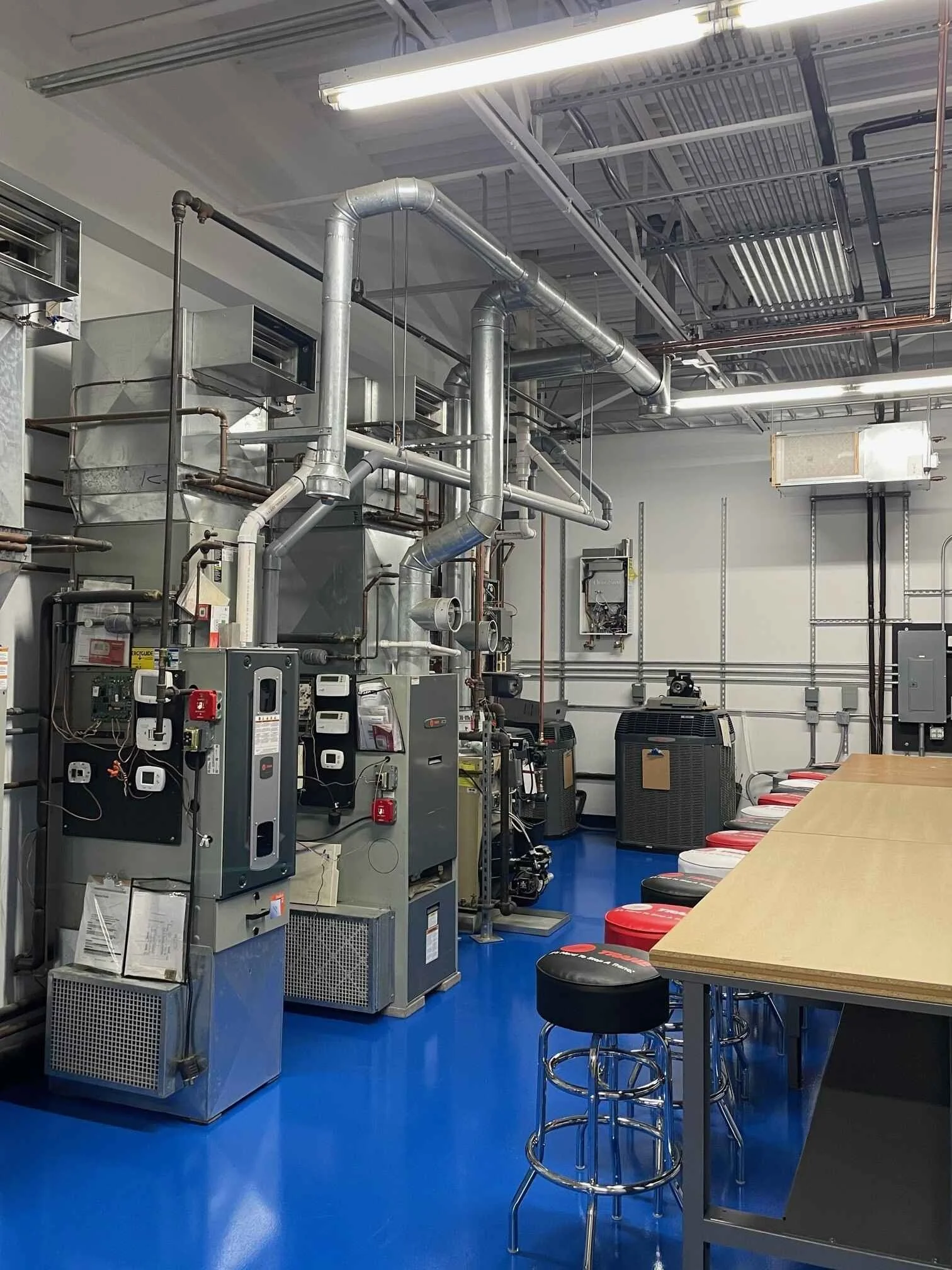 Industrial room with machinery, exposed pipes, and a blue floor, featuring a long wooden table with red, white, and black stools.