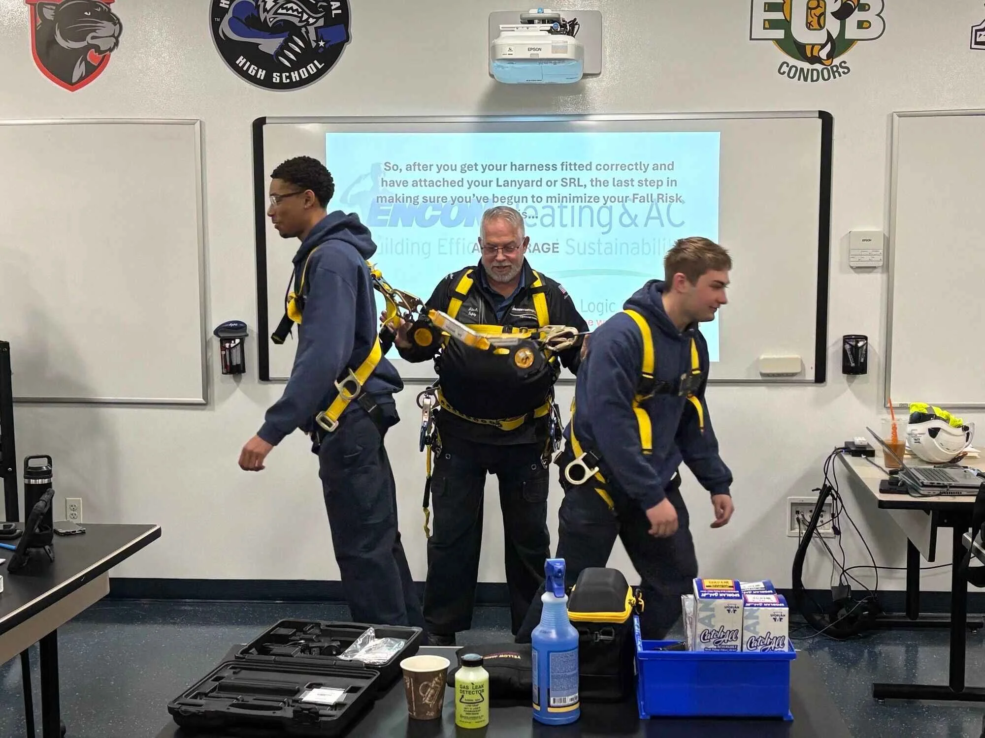 Three individuals wearing safety harnesses stand in a classroom setting, with a whiteboard and a projector screen behind them. The classroom has sports team logos on the wall and various safety equipment and supplies on the tables and shelves.
