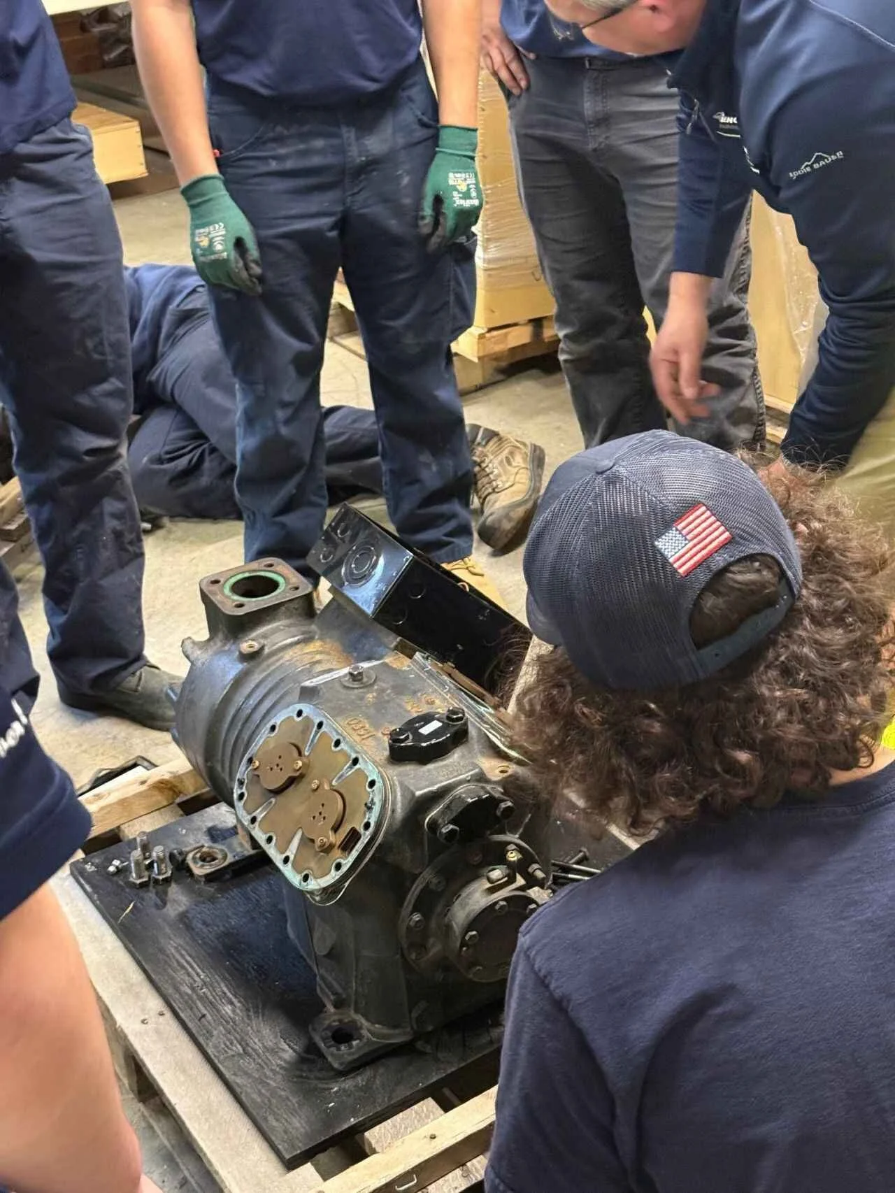 Group of people gathered around and examining a mechanical engine assembly on a table, in a workshop or garage setting.