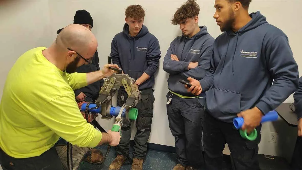 A man with a bald head, beard, and glasses demonstrates a plumbing fixture to four young men in a room. The young men are dressed in navy blue hoodies and black work pants, watching attentively. One of the young men holds blue and green pipe fittings