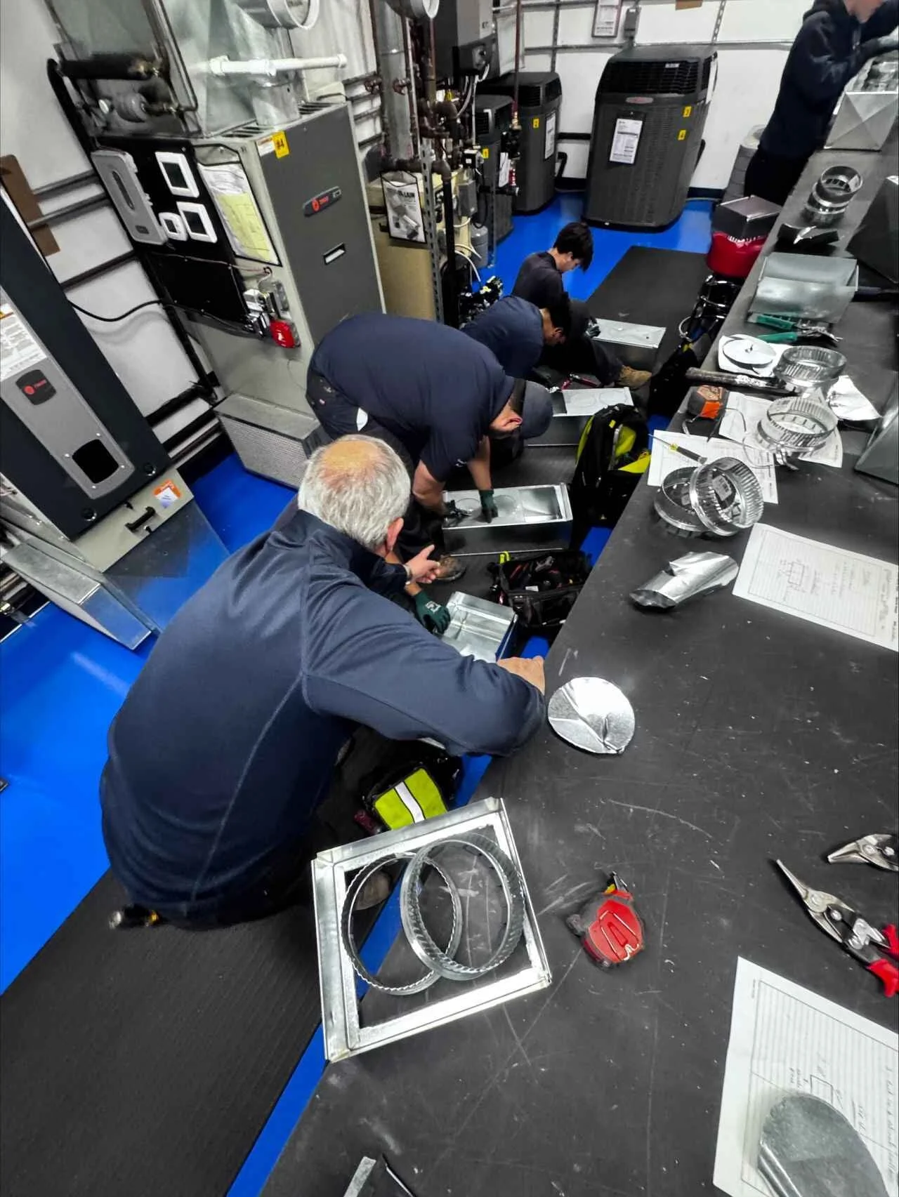 Group of people working on equipment in a workshop or industrial setting, with tools and metal parts on the table.