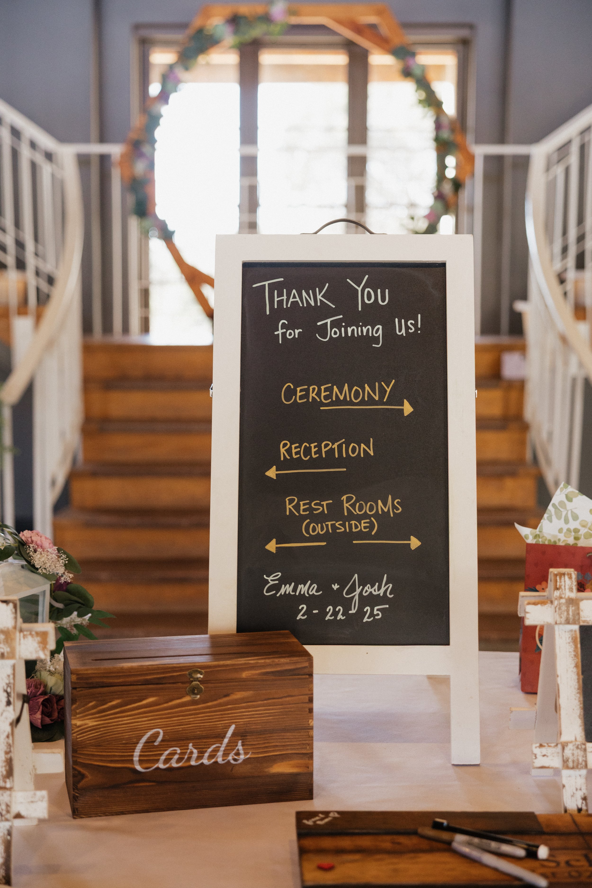 Wedding reception entrance with a chalkboard sign providing directions, a wooden card box labeled "Cards," and floral decorations. Stairs and a floral arch are in the background.