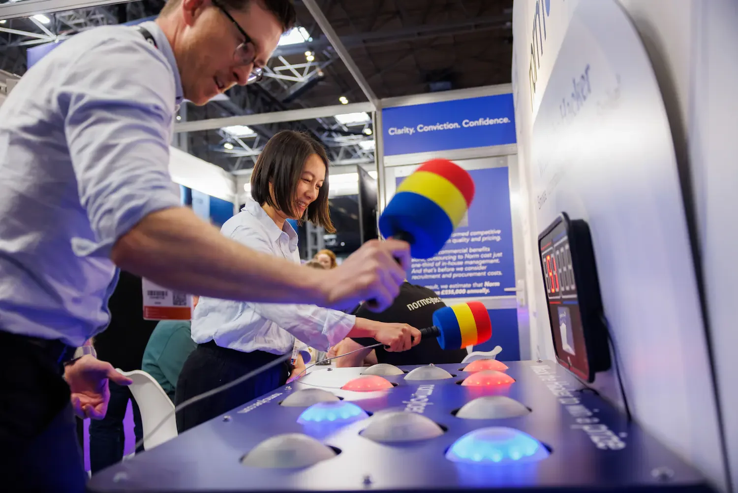 Two individuals, a man and a woman, playing a video game at a booth with large, colorful controllers attached to a screen, in a busy exhibition hall.