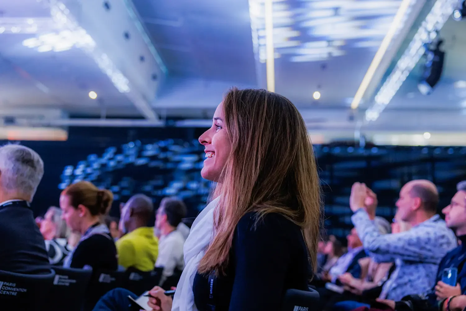 A woman with long brown hair smiling, sitting among an audience of people in a conference or event hall with blue and white lighting.