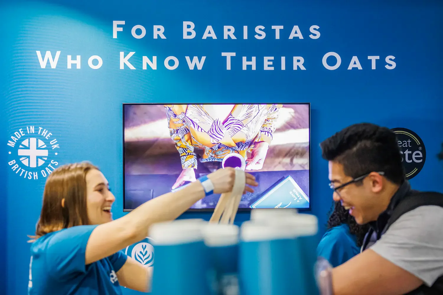 Two people smiling at a booth with a blue background that reads 'For Baristas Who Know Their Oats,' featuring a logo for 'Made in the UK British Oats' and 'Great Taste.' A woman and a man are interacting, with the woman holding a container and the ma
