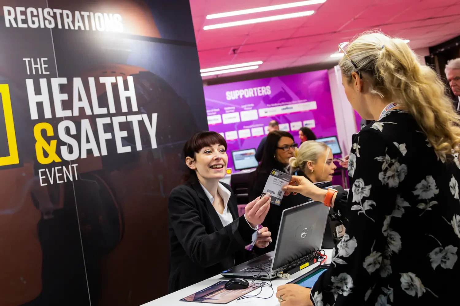 A woman at a registration desk handing a ticket to another woman during a health and safety event. Several people are visible in the background with computers and a purple "Supporters" wall.