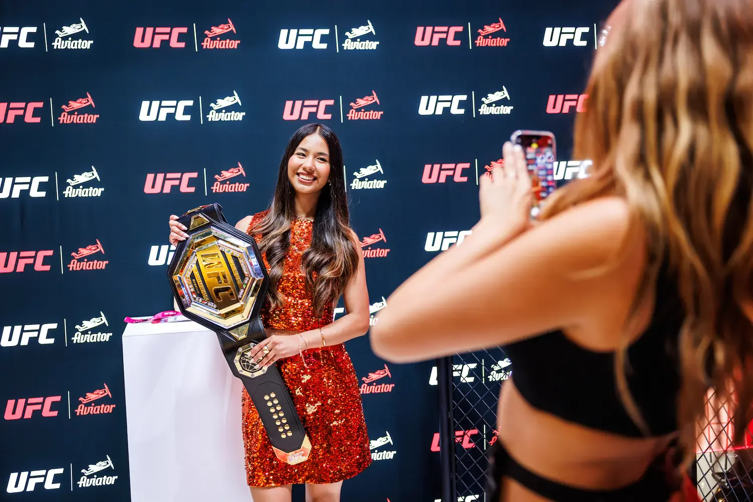 A woman in a red sequin dress holds a UFC championship belt and smiles while a woman with long hair in black attire takes a photo at a UFC event with a backdrop featuring UFC and Aviator logos.