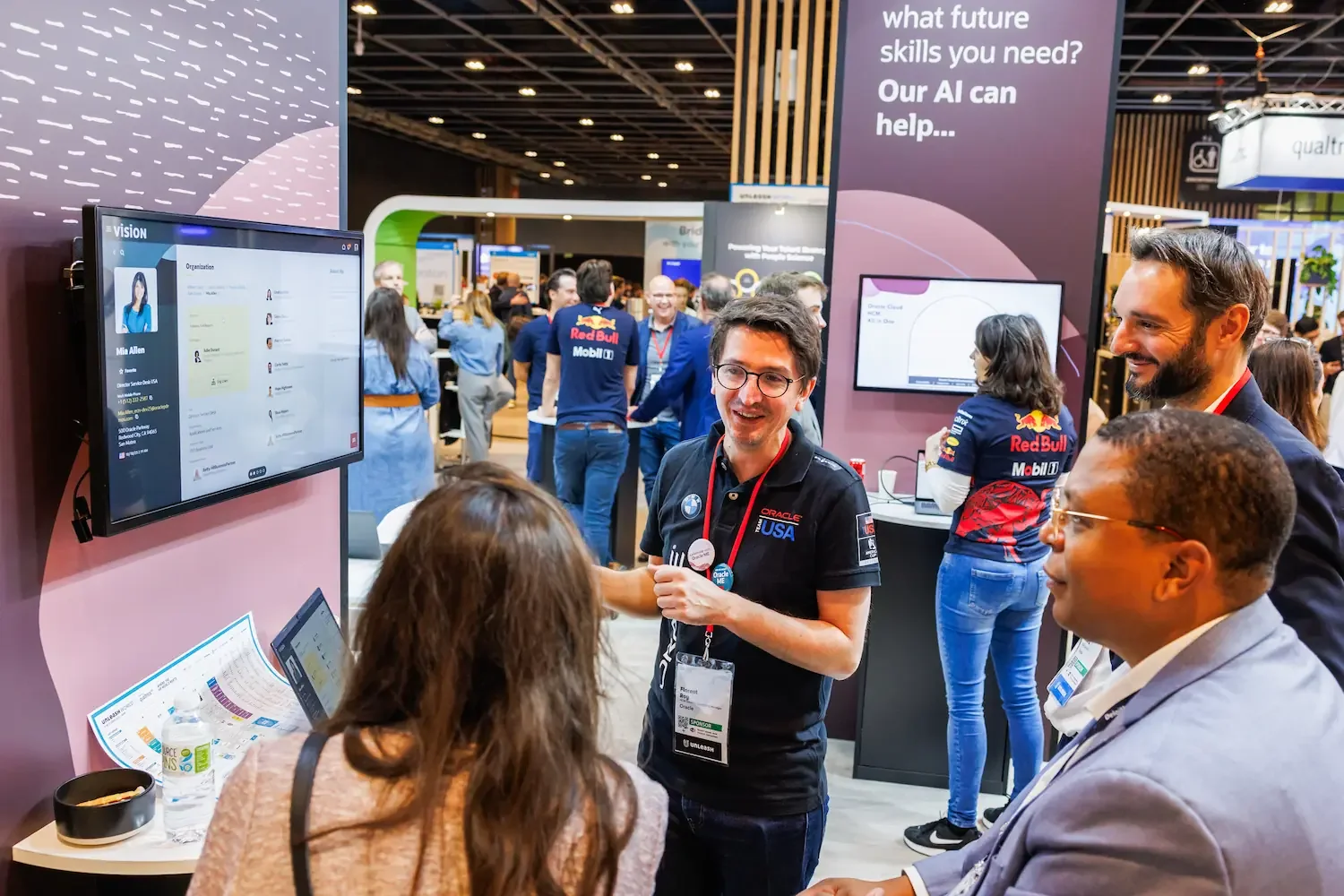 A group of people at a tech conference, with some engaged in conversation near a booth featuring screens displaying user profiles and digital content. The background shows a busy exhibition hall with other attendees and booths.