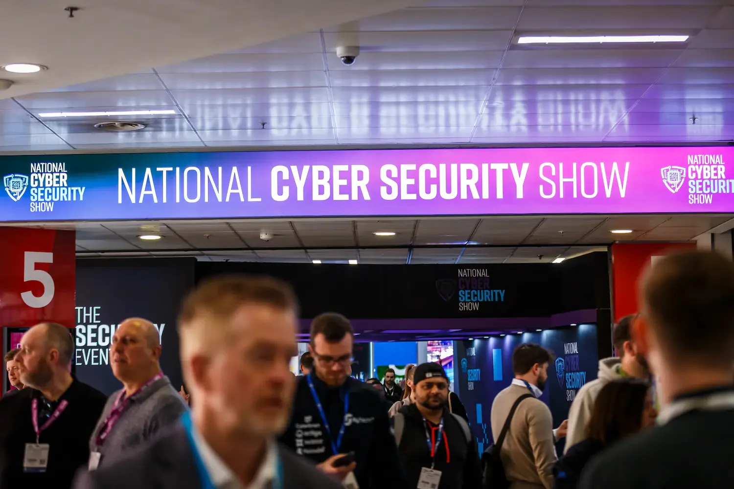 Entrance to the National Cyber Security Show with a purple and blue neon sign and conference attendees walking in.