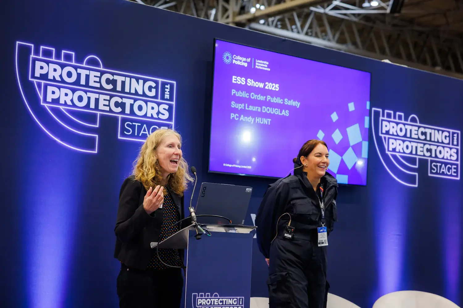 Two women on stage at a conference, smiling and speaking. Behind them is a large screen displaying event details, including 'ESS Show 2025' and topics about public order and public safety. The background has a blue theme with logos and the phrase 'Pr