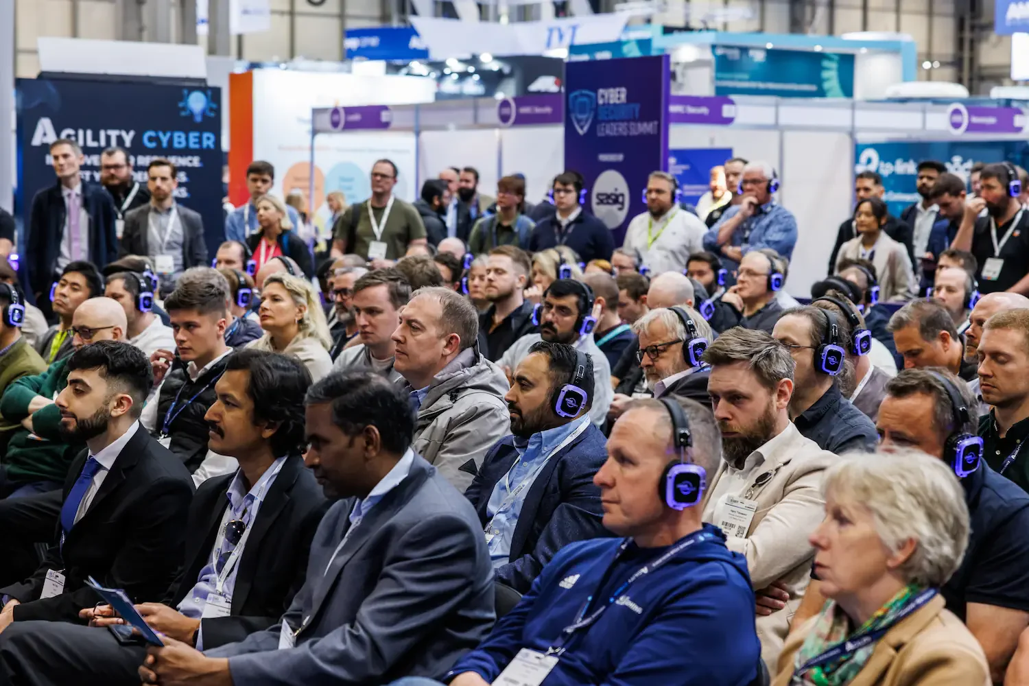 Audience attending a conference, many wearing blue headphones, sitting and listening to a speaker at a busy convention center.