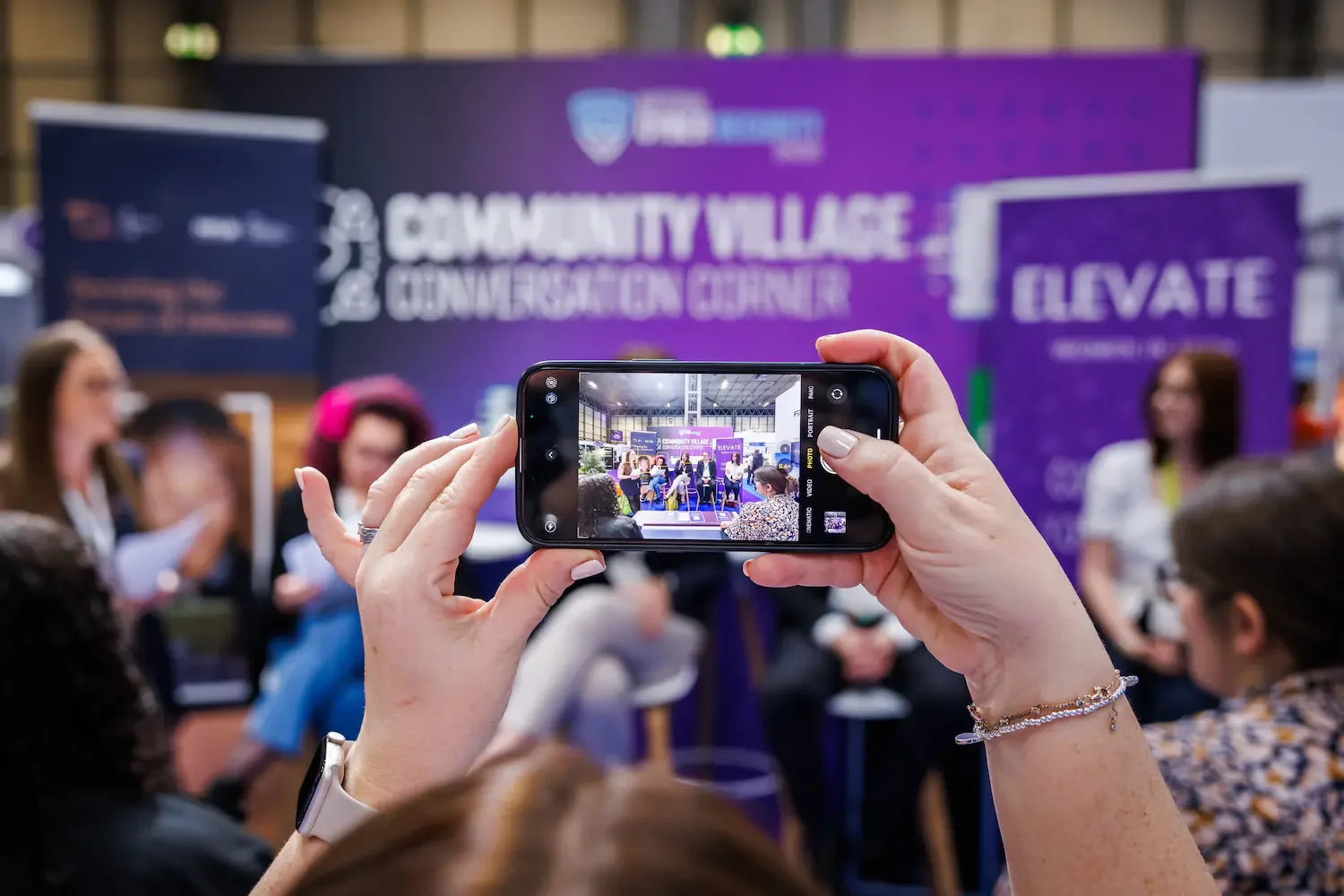 Person holding a smartphone taking a photo of a panel discussion at an event called Community Village Conference Center, with several people seated on the stage.