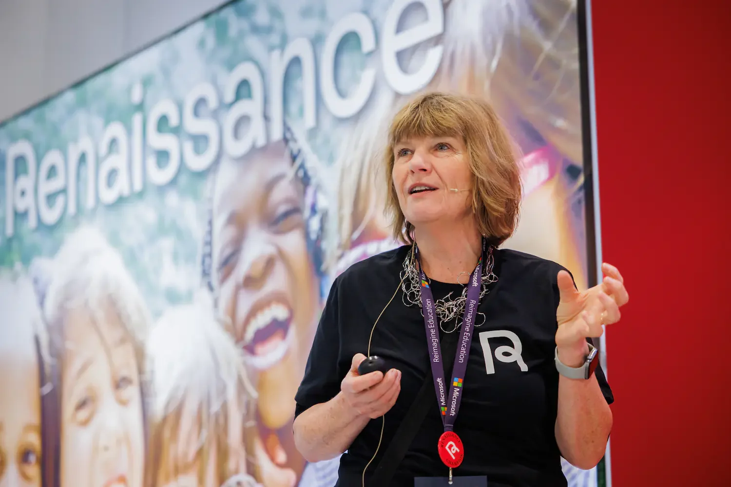 A woman with shoulder-length light brown hair giving a presentation in front of a large screen displaying the word 'resilience' and a smiling child's face. She is wearing a black T-shirt with a logo, a purple lanyard, and a headset microphone.