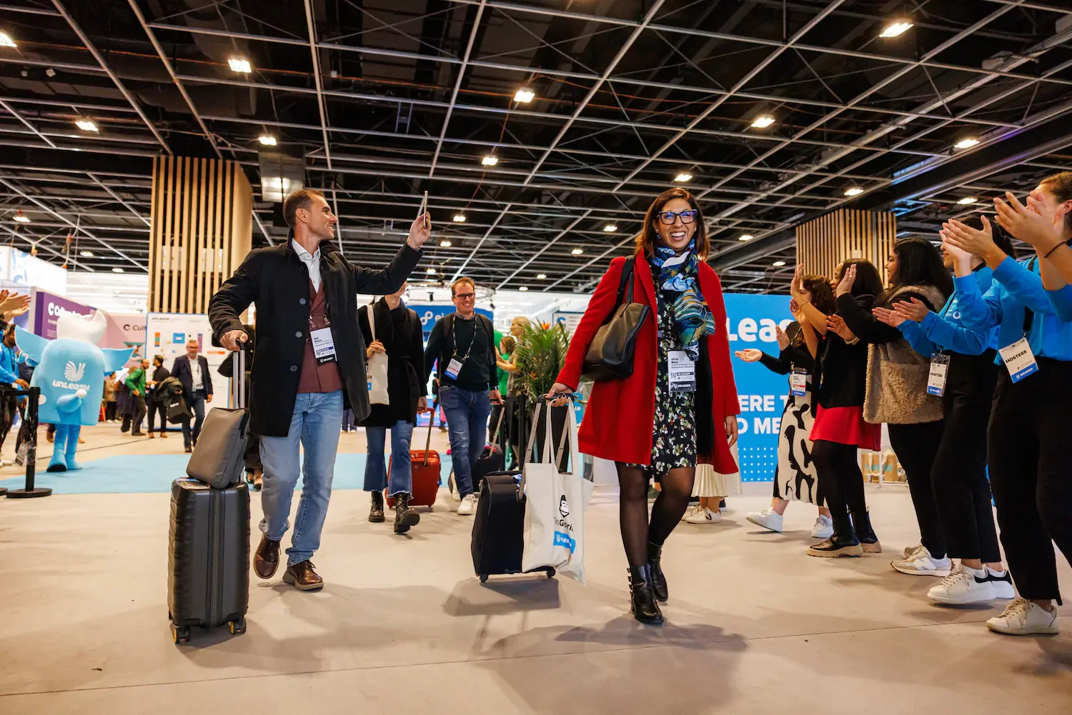 People arriving at an airport with luggage, walking through a welcoming crowd with smiles, in a large indoor space with high ceilings and bright lighting.