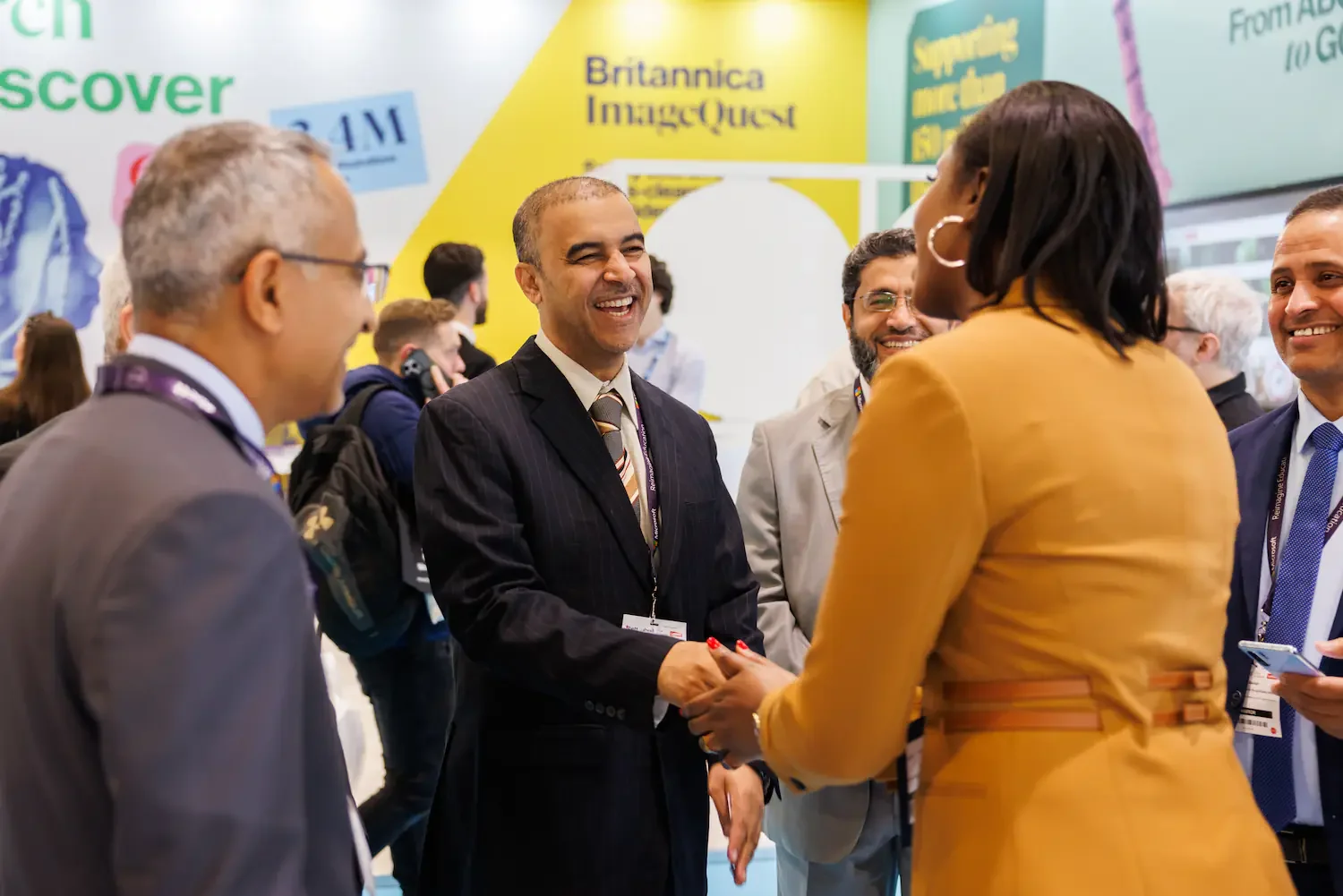 Group of diverse professionals at a conference, smiling and engaging in conversation, with a colorful backdrop featuring the Britannica ImageQuest logo and various text signs.
