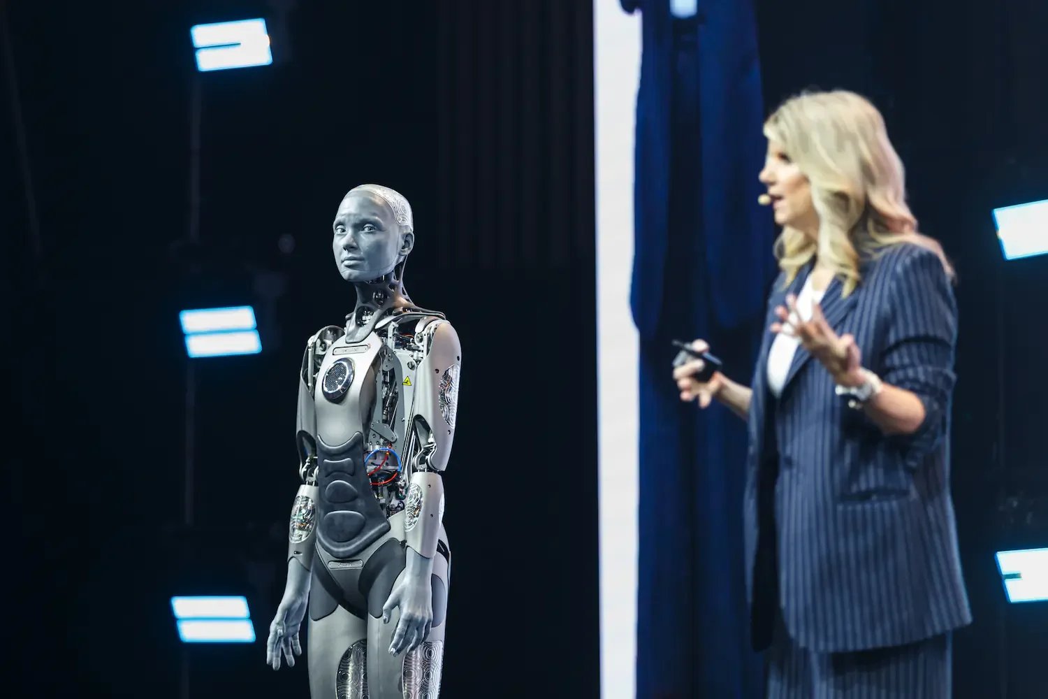 A woman giving a presentation next to a humanoid robot on stage, with stage lights in the background.