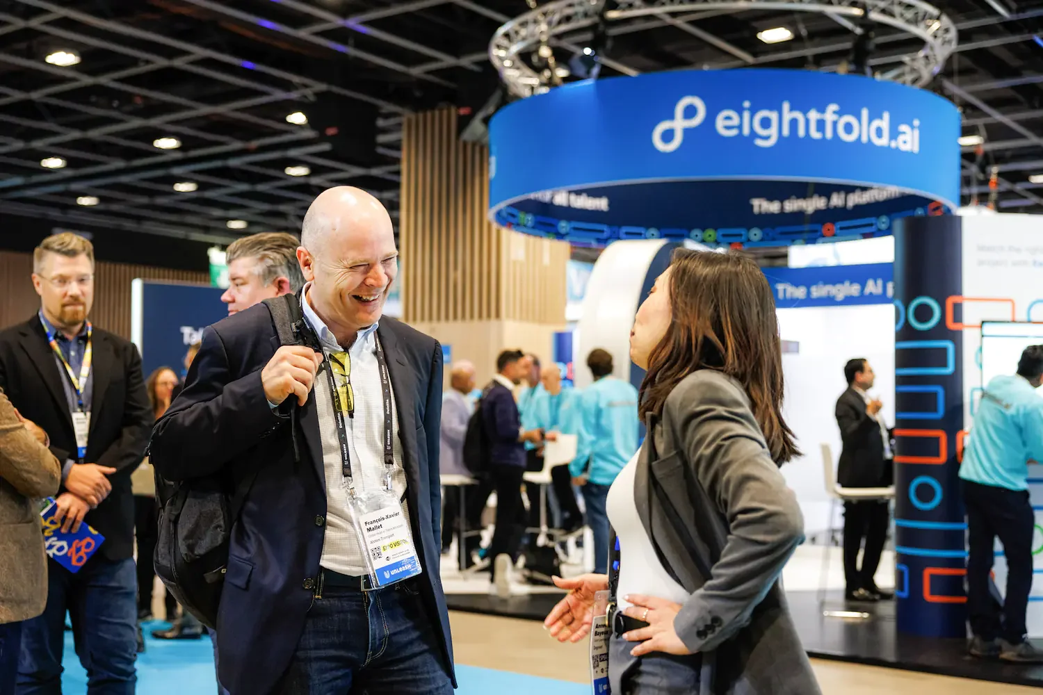Two business professionals, a man and a woman, are engaged in conversation at a trade show or conference. The man is smiling and holding a backpack strap, while the woman is gesturing with her hand. In the background, there are other attendees and a 