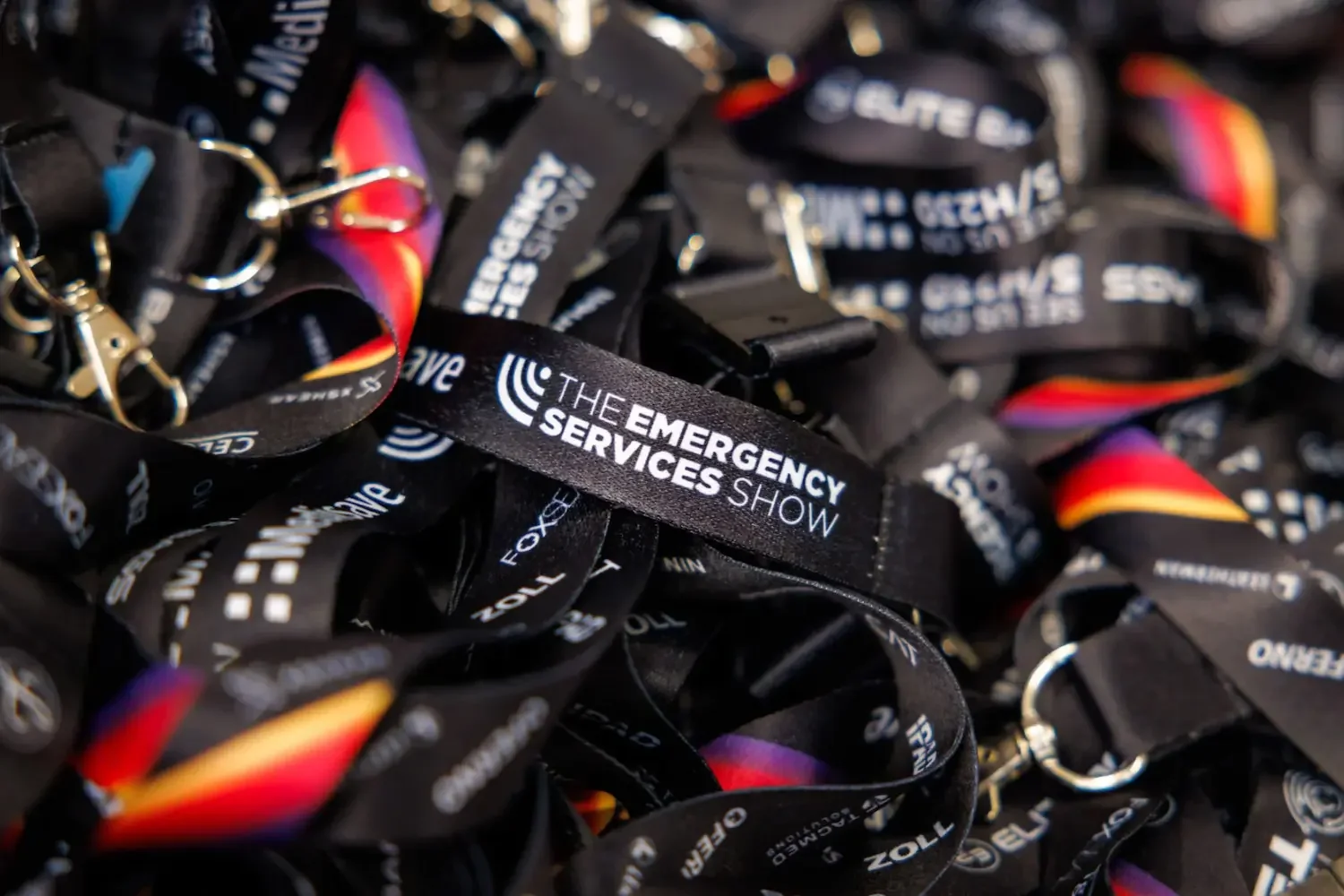 A pile of black lanyards with white text that read "The Emergency Services Show" and a colorful rainbow stripe on some of them.