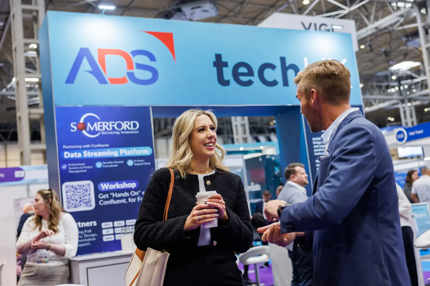 A woman and a man are having a conversation at a tech conference. The woman is smiling and holding a coffee cup, wearing a black blazer with a white top. The man is wearing a blue blazer and is gesturing with his hands. The background shows a large b