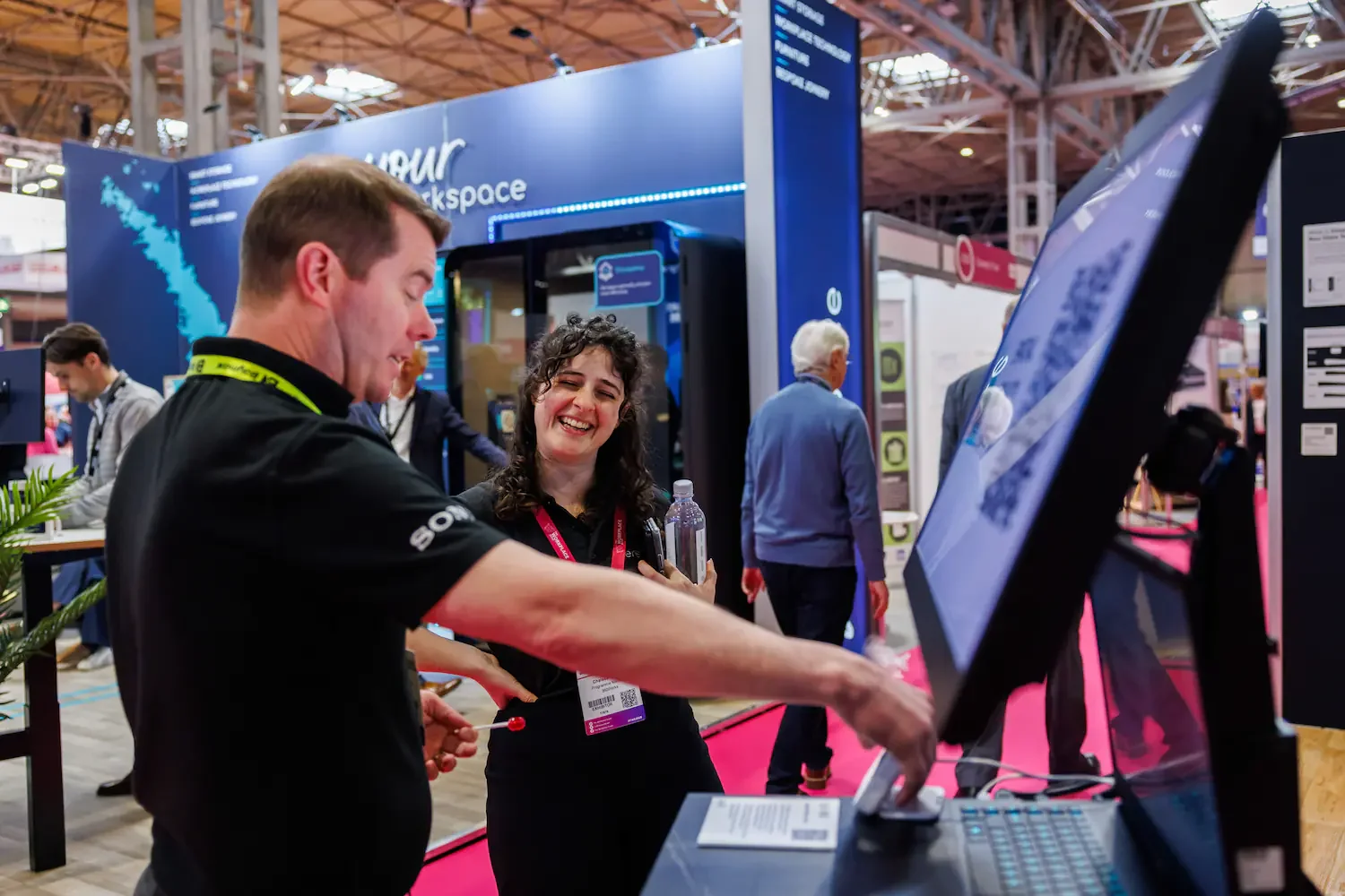 Two people at a technology expo, one showing a feature on a computer screen, smiling and engaging with each other. The background shows other attendees and booths with informational displays.