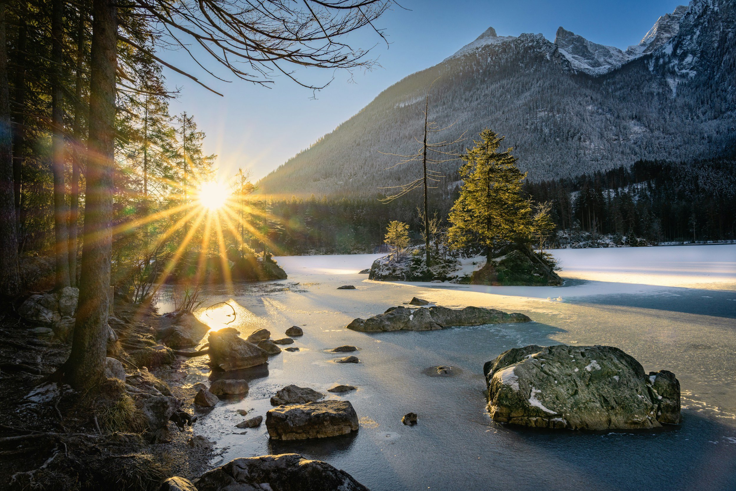 Sunset over a partially frozen lake surrounded by trees and mountains.
