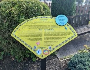 An informational sign in a garden, shaped like a leaf, containing educational content about bees and biodiversity, surrounded by plants and a small bench.