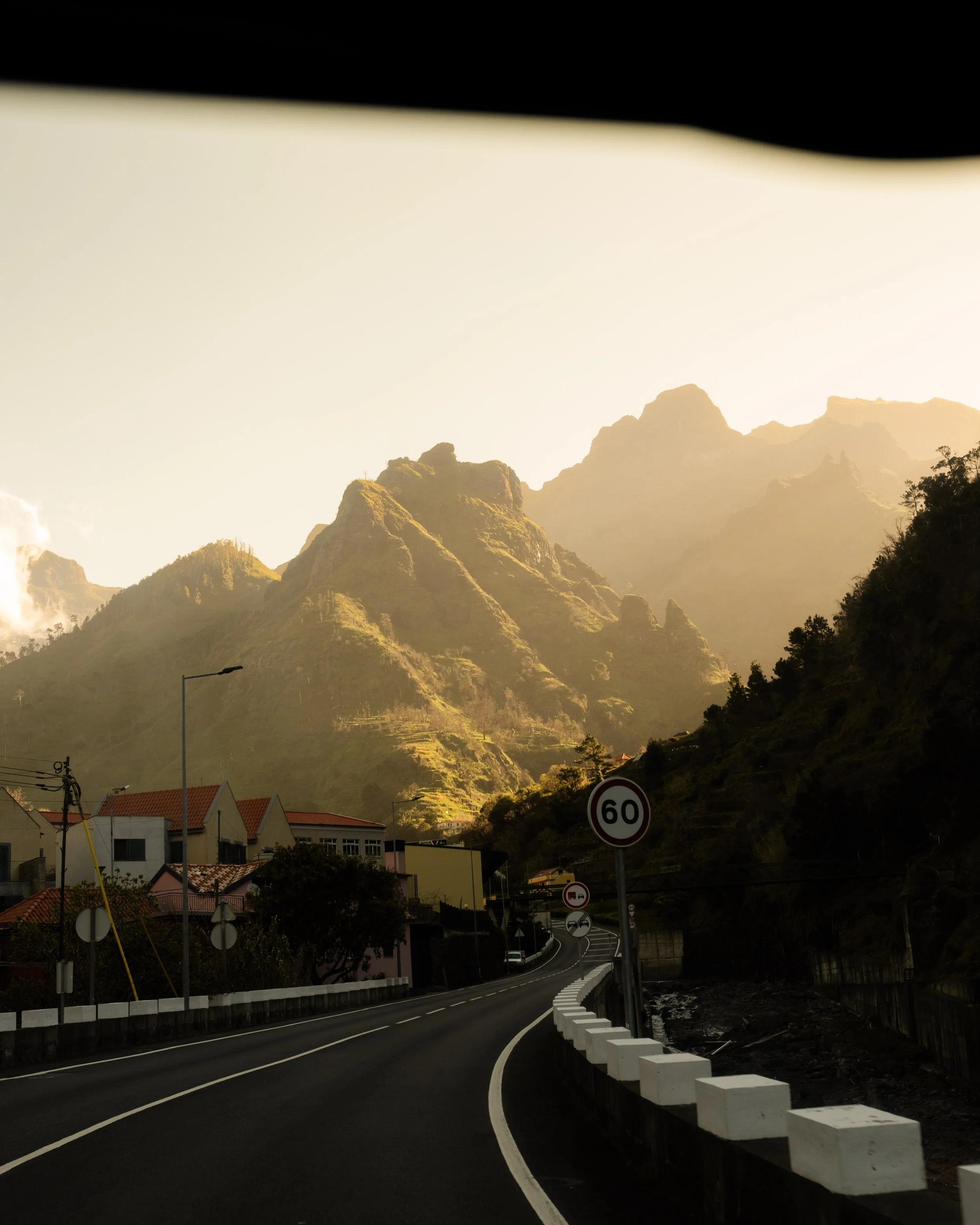 A mountain landscape with a winding road in the foreground, residential houses with red-tiled roofs, street signs, and a mountain range in the background during sunset.